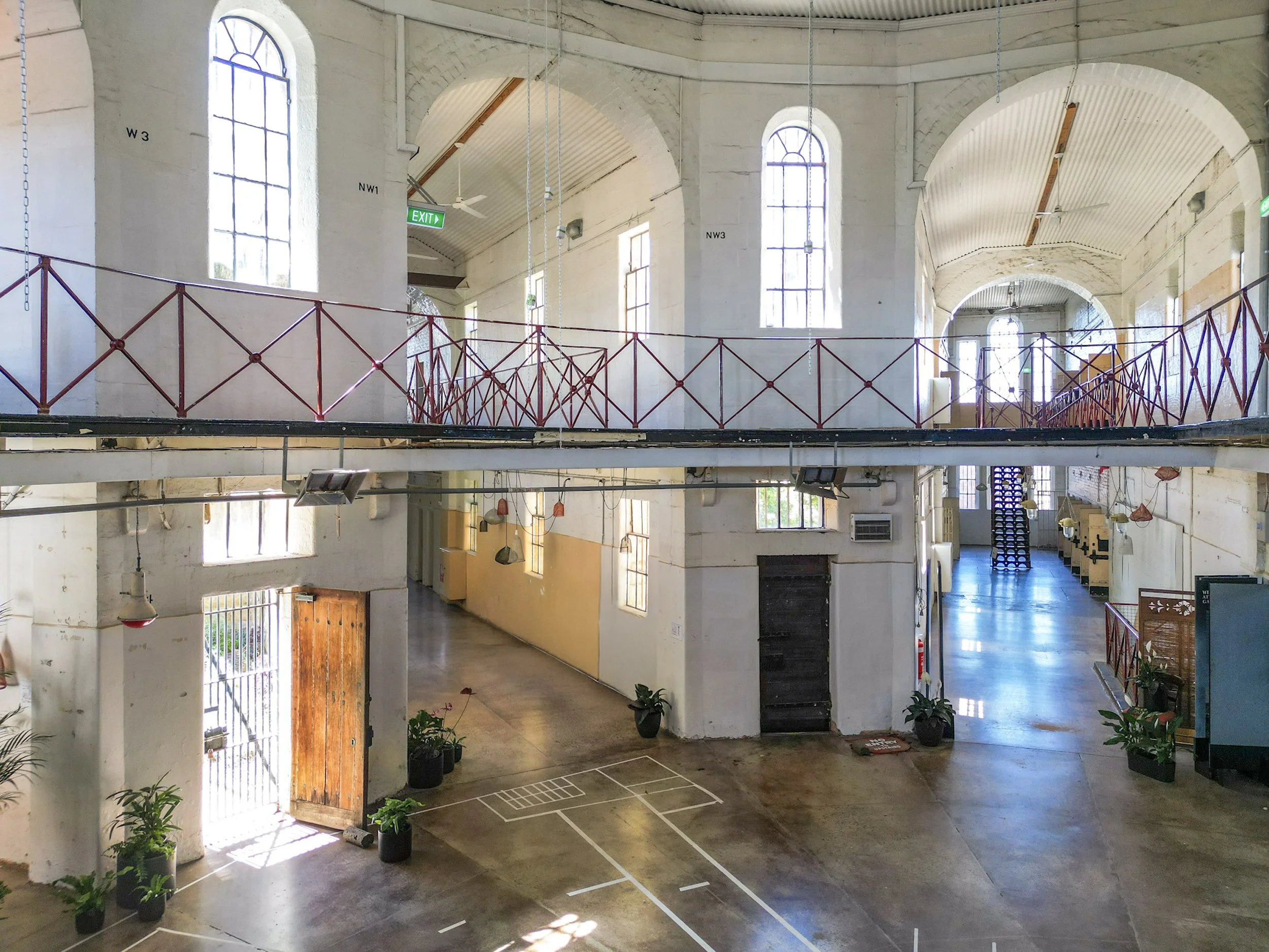 Interior of a spacious, high-ceilinged building with large arched windows, a second-floor balcony with a red railing, hanging lights, and plants on the ground floor.