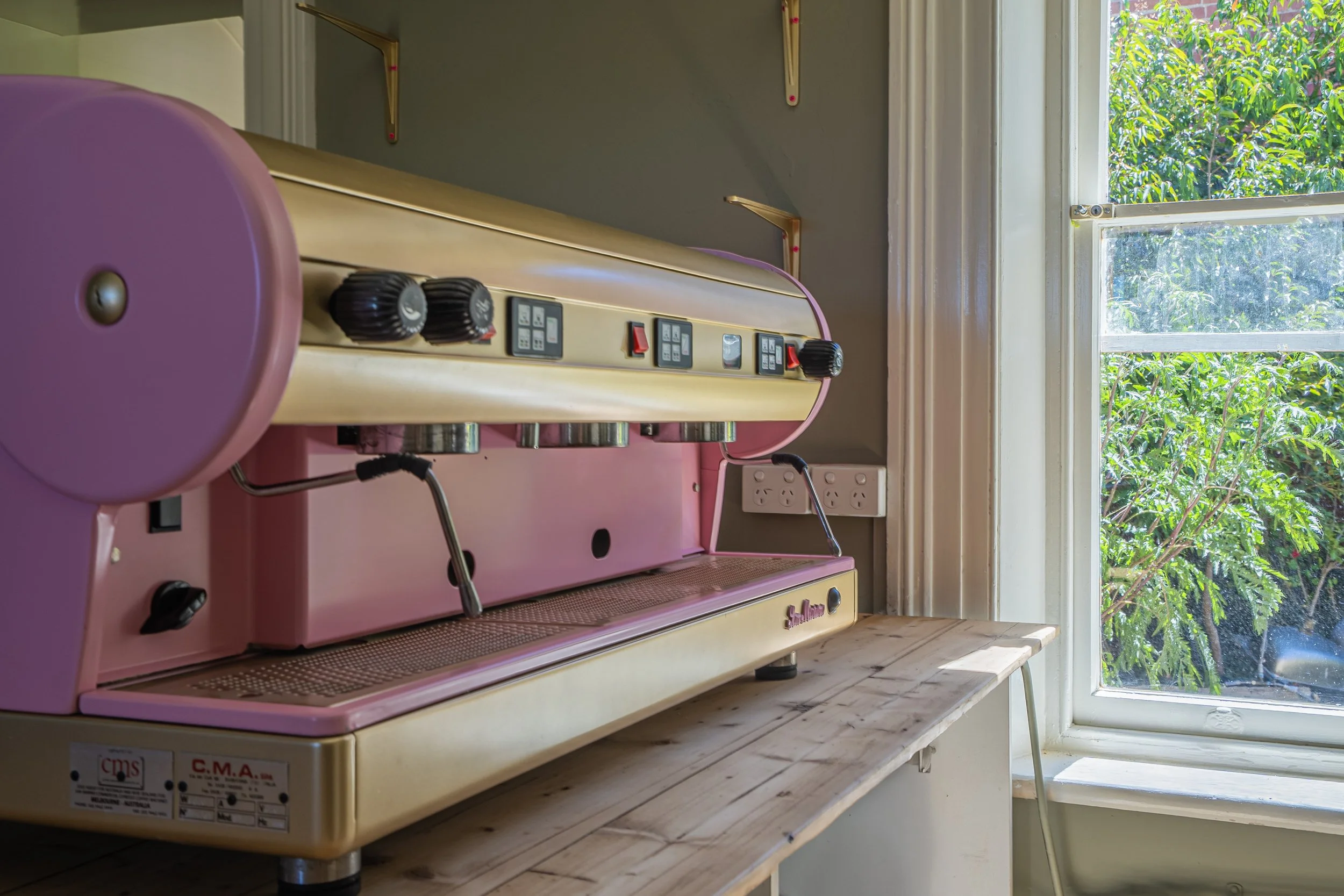 A pink and cream vintage espresso machine on a wooden countertop near a window with green trees outside.