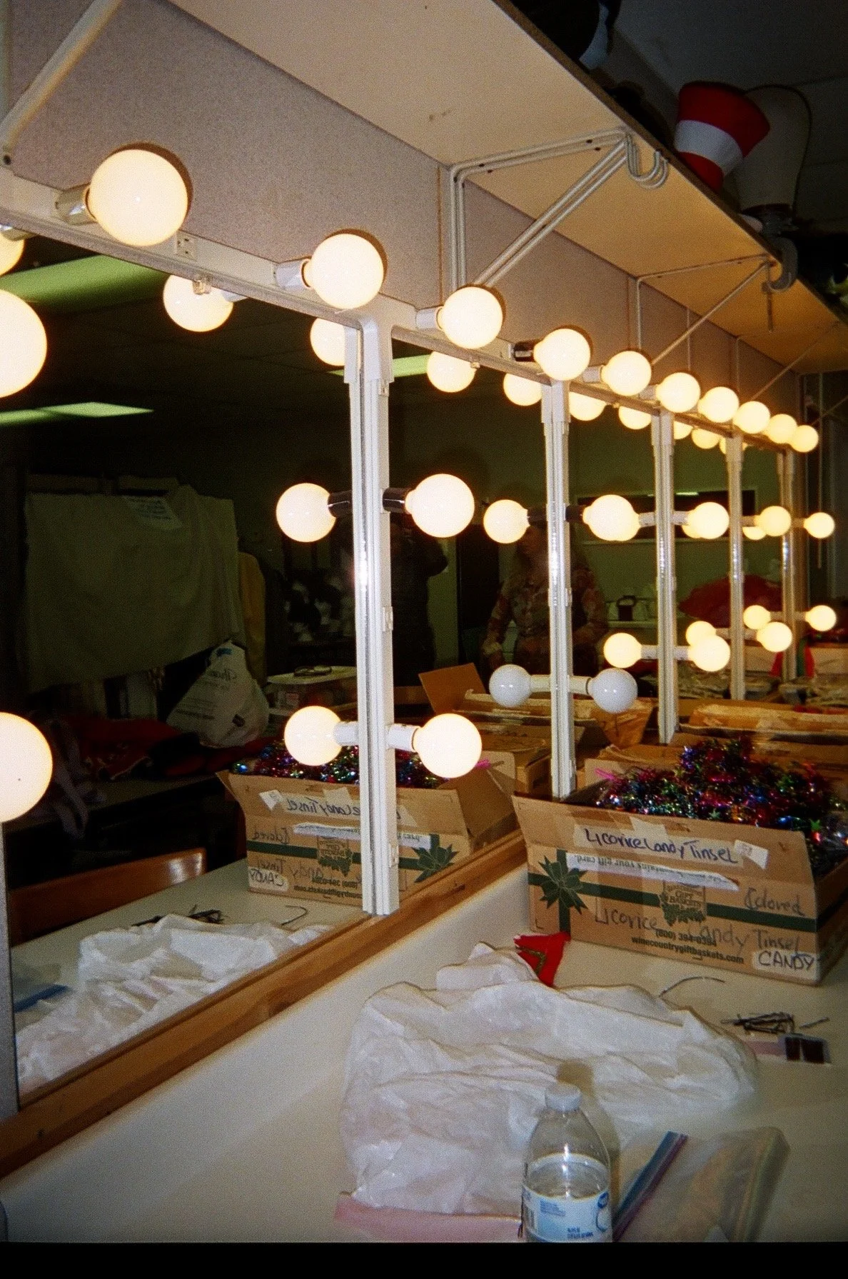 Elana Kline Art Photography: Mirror with light bulbs, cardboard boxes filled with Christmas decorations, a water bottle, and some tissue paper on a desk or table.
