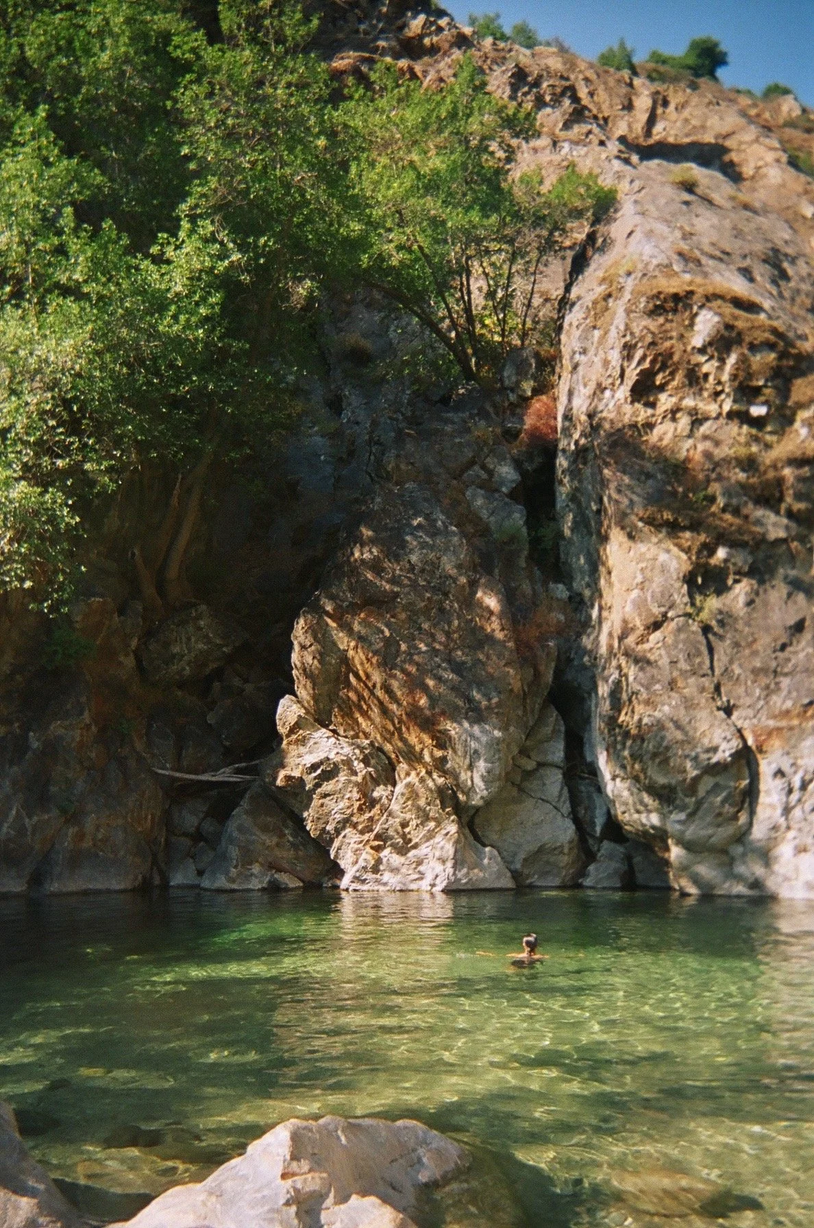 Elana Kline Art Photography: A person swimming in a natural water pool surrounded by large rocks and green trees, with a hillside in the background.