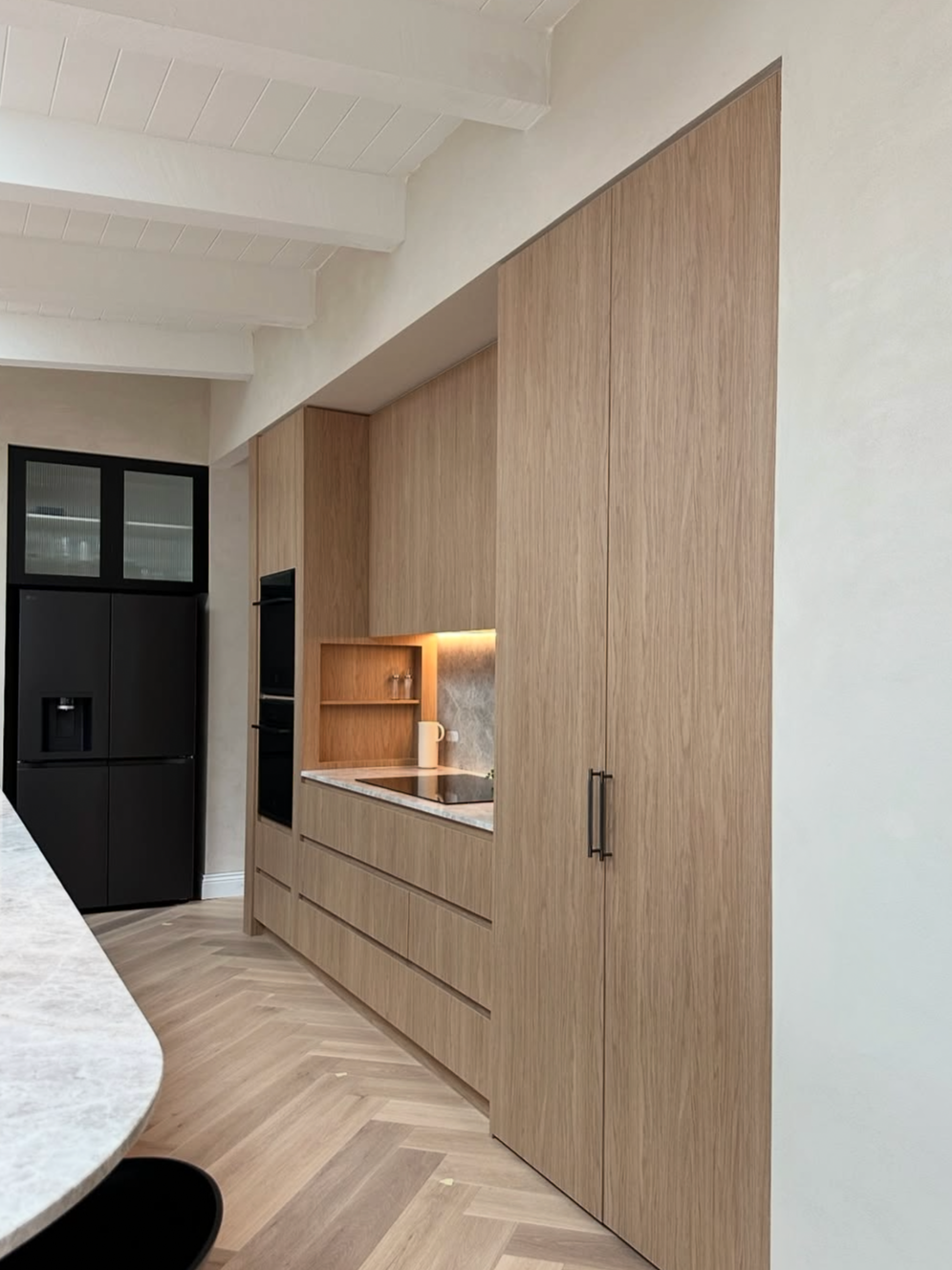 A modern kitchen with light wood cabinetry, a built-in oven, a small open shelf with glasses, and a black refrigerator.