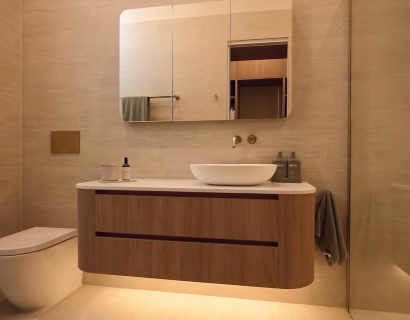 Modern bathroom with a floating wooden vanity, white vessel sink, wall-mounted gold faucet, and a mirrored cabinet. Beige tiled walls and a reflected doorway with wood paneling. Items on the vanity include a black bottle, a gray bottle, and a small container. A towel hangs on a hook to the right.