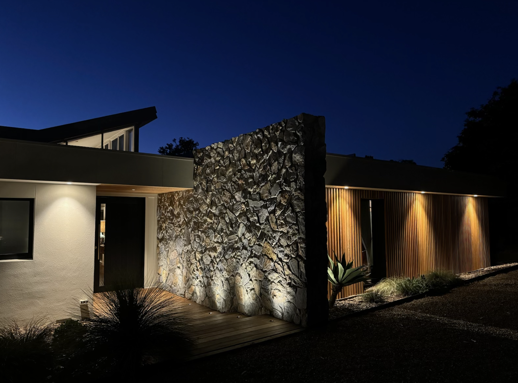 A modern house at night featuring a textured stone wall, lighting, garden lighting on stone. High end lighting on stone.