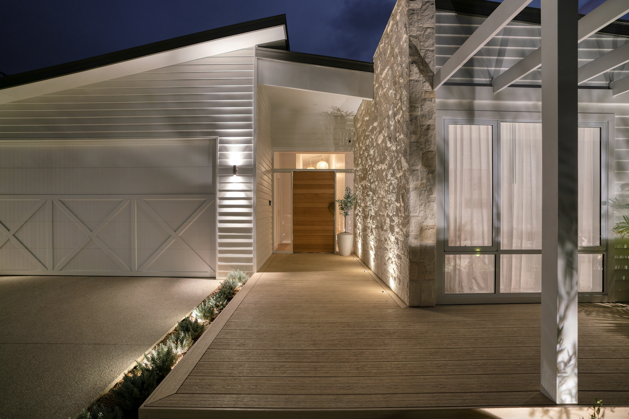 Night view of a modern house entrance with a wooden door, stone wall, and white siding, illuminated by exterior lighting, with a wooden deck and small plants along the side.