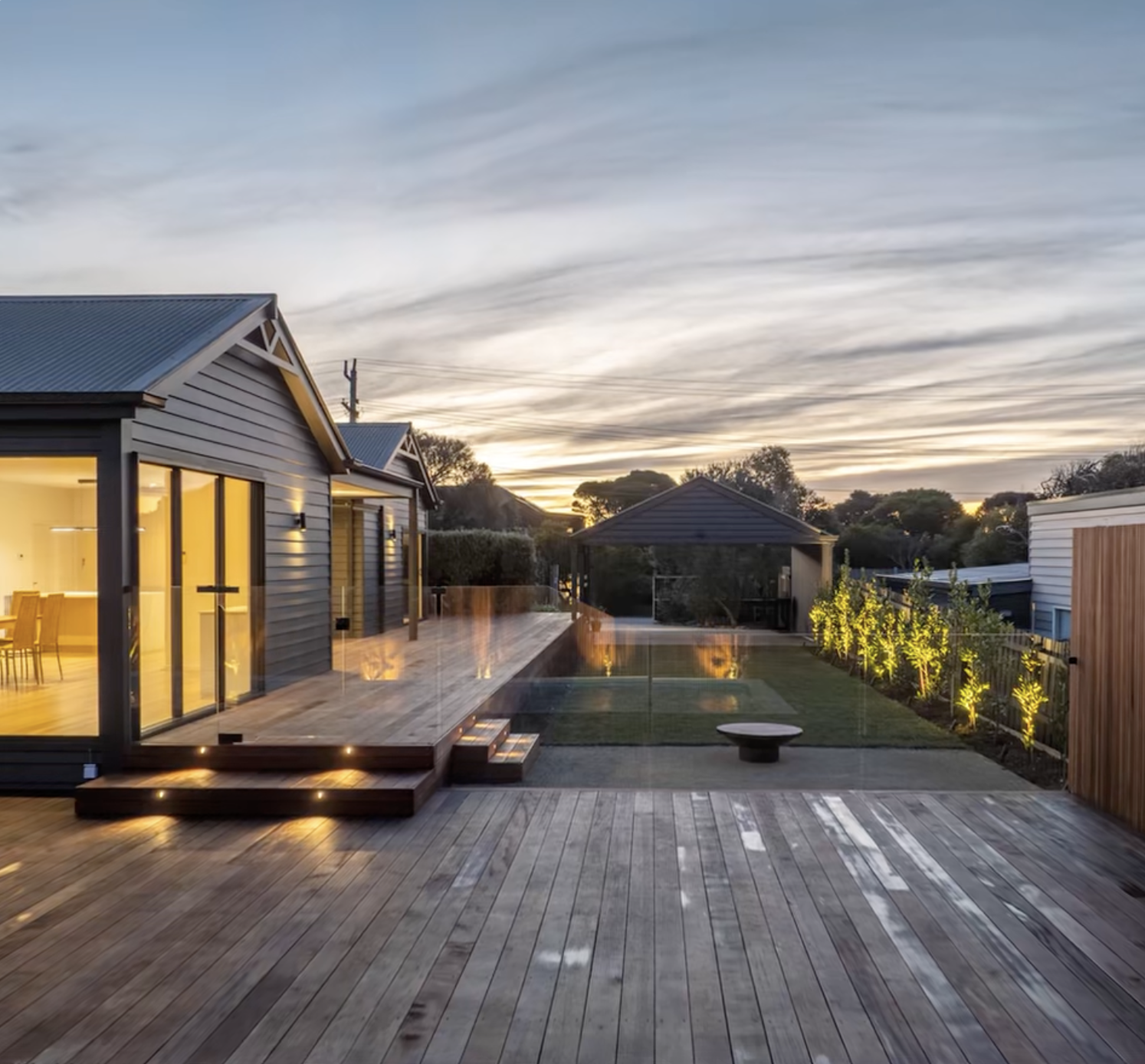 A modern backyard patio with wooden decking, a small grass area, and a fire pit. The house has large sliding glass doors with lights on inside, and there are small illuminated trees along the fence on the right side. The sky is dusky with scattered clouds.