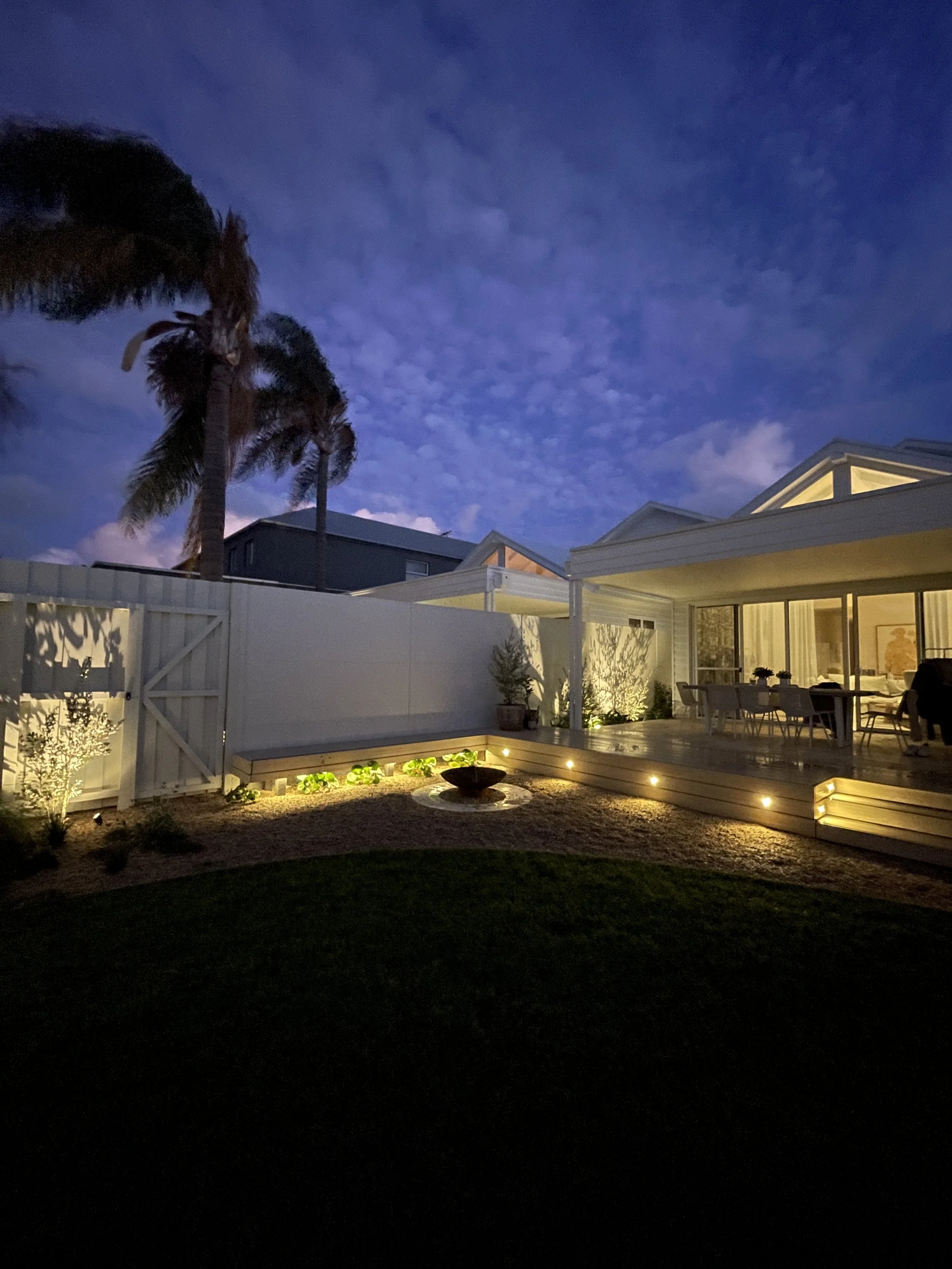 A backyard patio at night with outdoor lighting, a deck with dining furniture, a small fountain, palm trees, and a house with large windows showing the lit interior, under a cloudy sky.