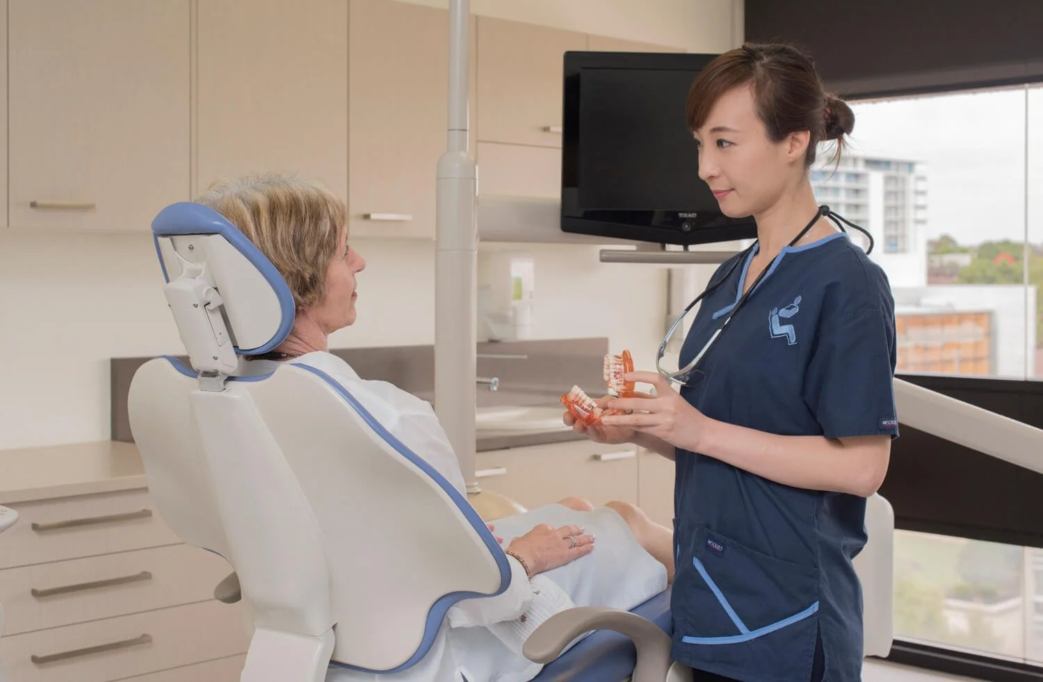 A young female nurse showing orthodontic dentures to an elderly woman sitting in a dental chair in a dental office.