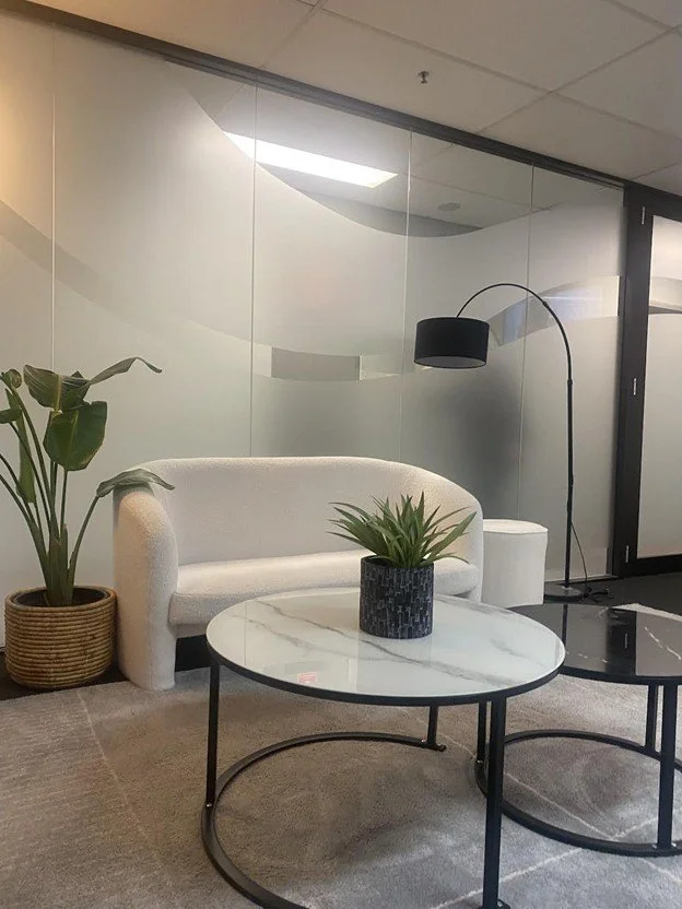 Modern office waiting area with a white sofa, a black floor lamp, a small white side table, a marble-top coffee table with a potted plant, and a large potted plant in a woven basket. The area has a glass wall with frosted design and a gray carpeted f