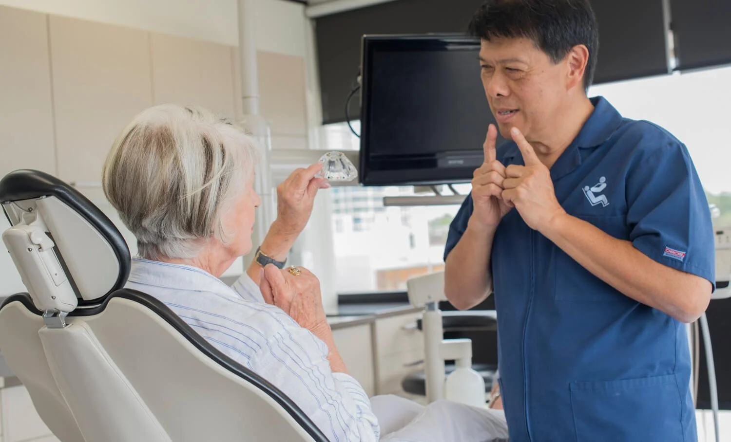 A healthcare professional and an elderly woman in a hospital room. The healthcare worker is wearing scrubs and appears to be in a playful or lighthearted moment with the woman, who is sitting in a hospital bed.