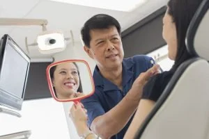 A dentist examining a patient's mouth with the reflection of a smiling woman in a mirror.