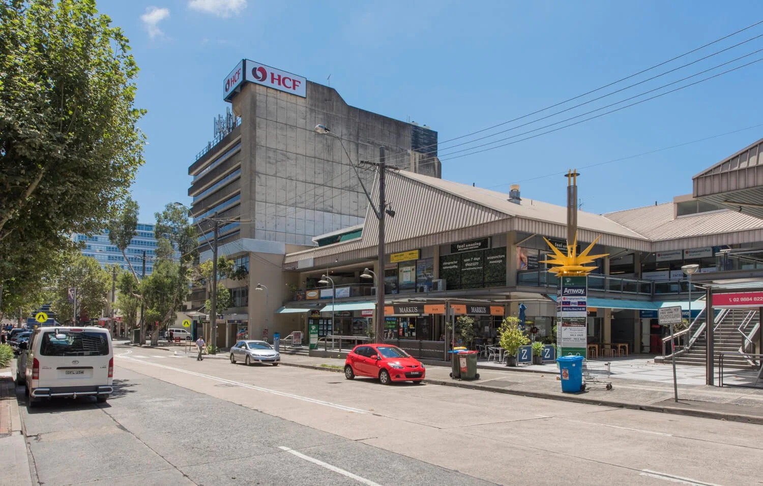 City street scene with parking cars, trees, buildings including a multi-story structure with 'HCF' signs, and a pedestrian walking.