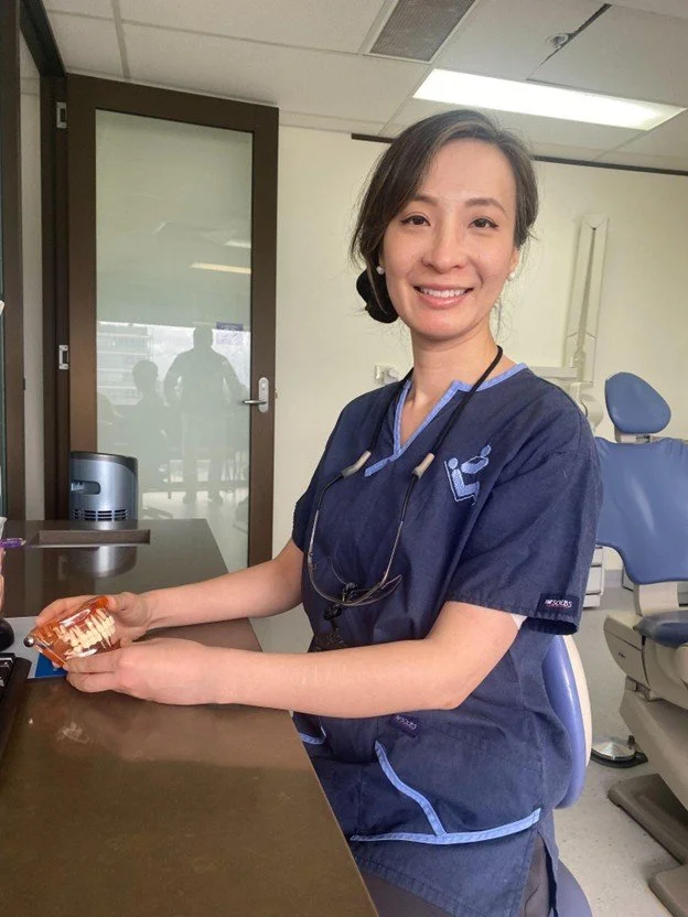 A smiling female healthcare professional wearing navy scrubs and stethoscope sitting at a desk in a medical office, holding an object in her hands.