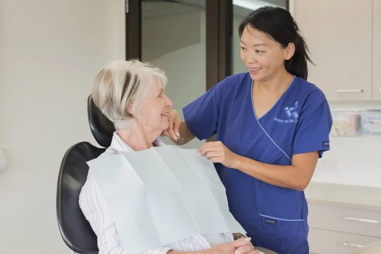A nurse in blue scrubs smiling and talking with an elderly woman sitting in a dental chair in a clinical setting.