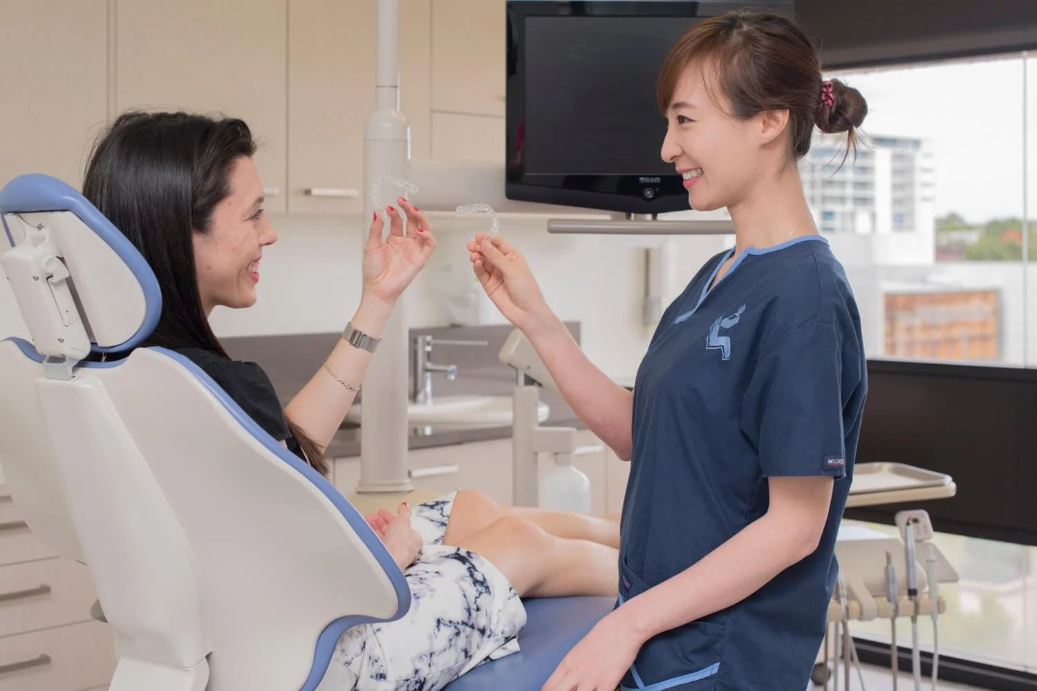 A woman in a dental chair smiling and holding a clear dental aligner while a dental professional, standing next to her, gestures towards another aligner. The setting is a modern dental clinic.