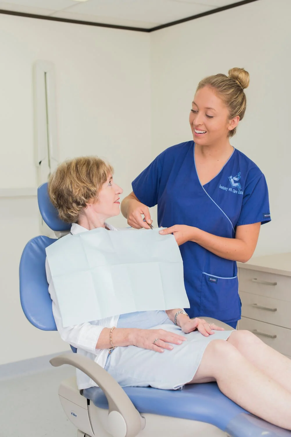 A nurse in blue scrubs administers an injection to a woman patient who is sitting in a dental chair, looking at the nurse and smiling.