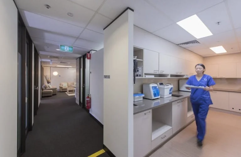 Healthcare worker in blue scrubs walking through medical or hospital kitchen area with white cabinets and medical equipment.