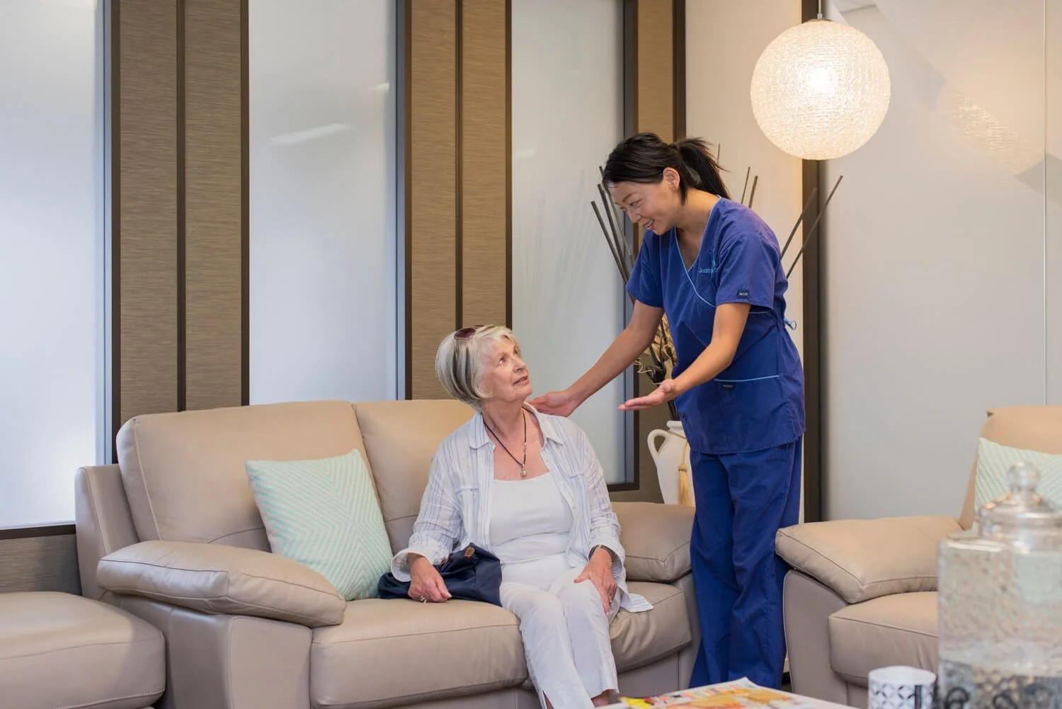 A nurse in blue scrubs talking to an elderly woman sitting on a beige sofa in a well-lit room.
