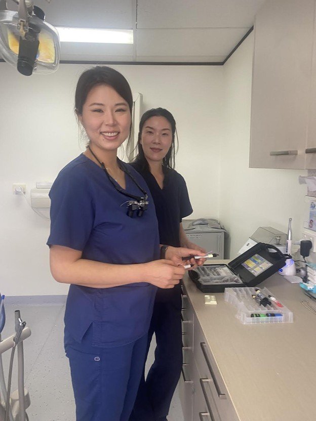 Two female healthcare professionals in scrubs standing in a medical room, smiling at the camera. One is holding a small medical device, with medical supplies and equipment visible on the counter.