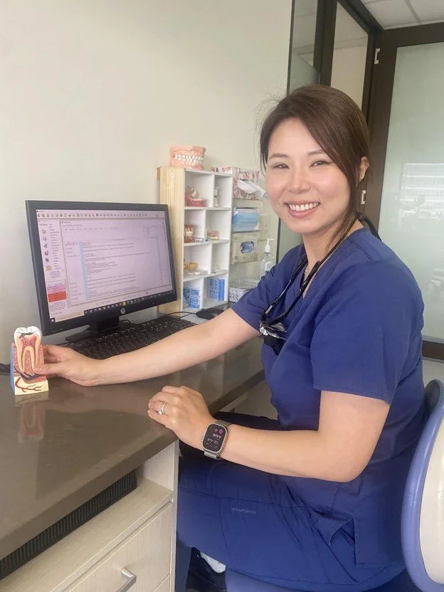 A female healthcare professional in blue scrubs sitting at a desk, smiling, with a computer and a dental model on the table, in a medical office or clinic.