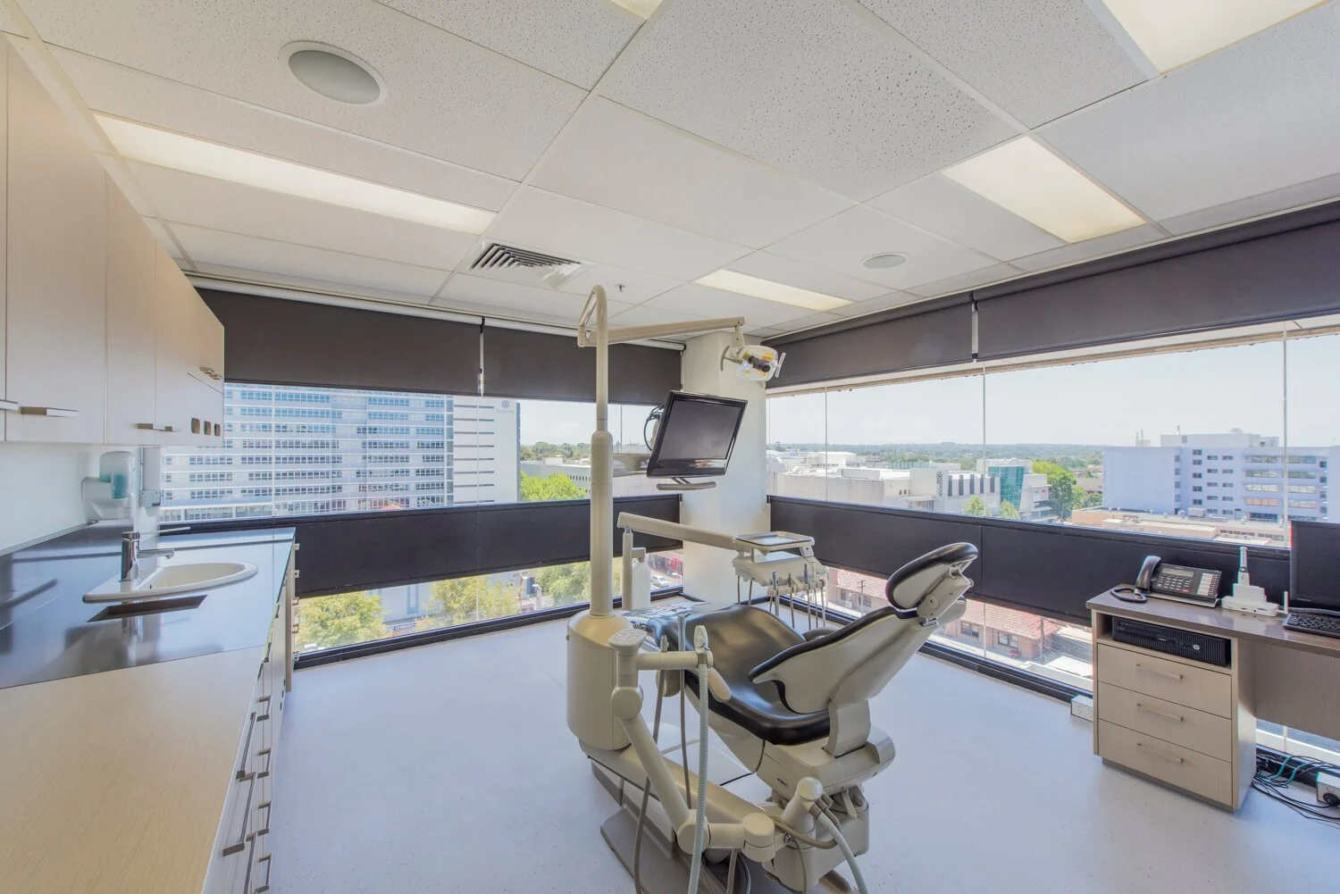 A dentist's office with a dental chair, a monitor, a dental tool, a cabinet with a phone and office supplies, and large windows showing a cityscape outside.