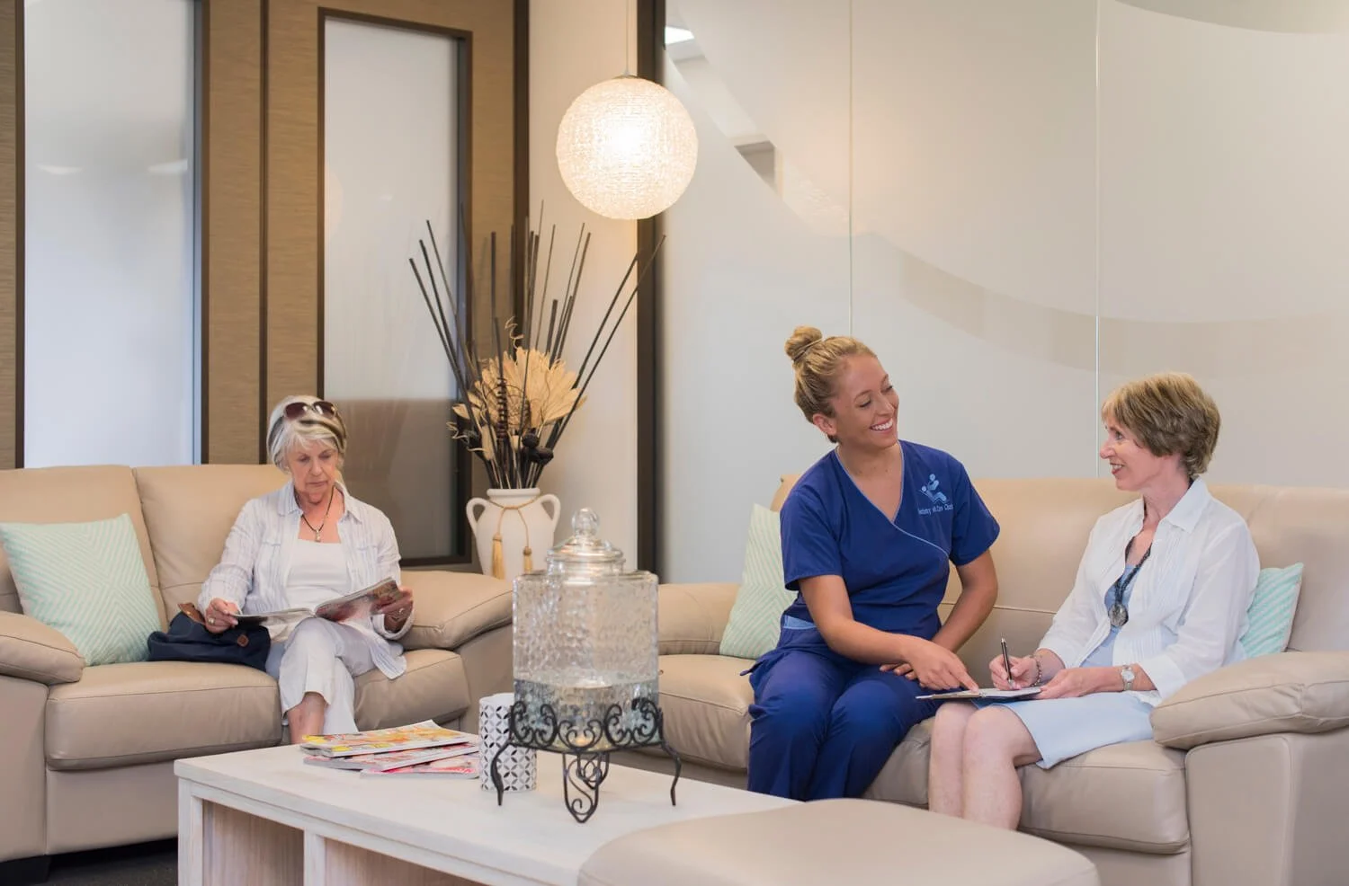 Three women sitting on a beige couch in a waiting room, two are having a conversation while the third is reading a magazine.