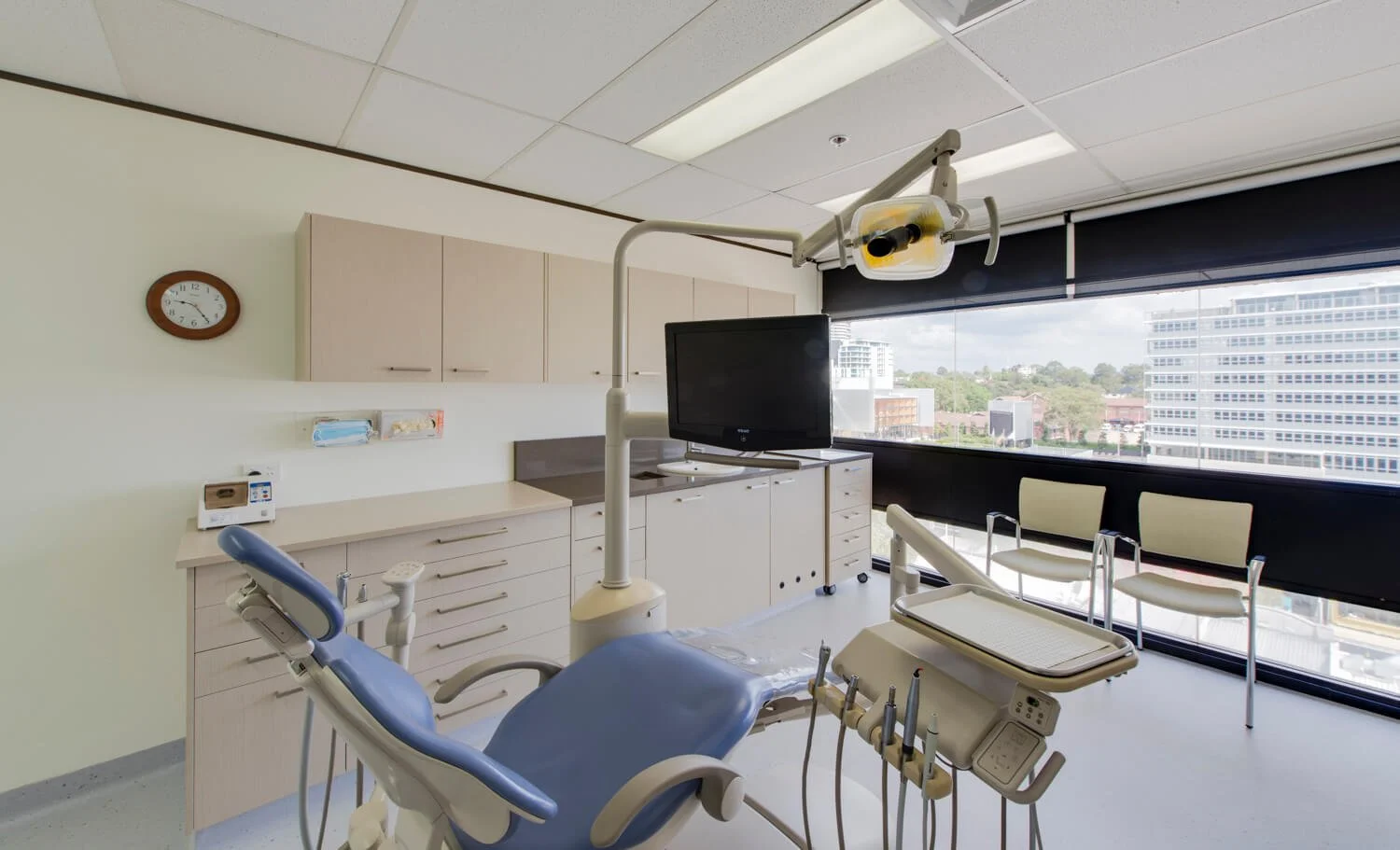 Empty dental examination room with a dental chair, overhead light, monitor, window view of city buildings, and chairs for visitors.