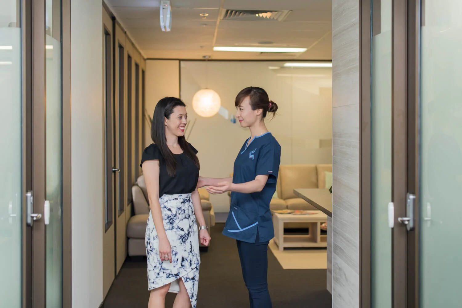 A nurse and a woman having a conversation in a medical clinic or office hallway.