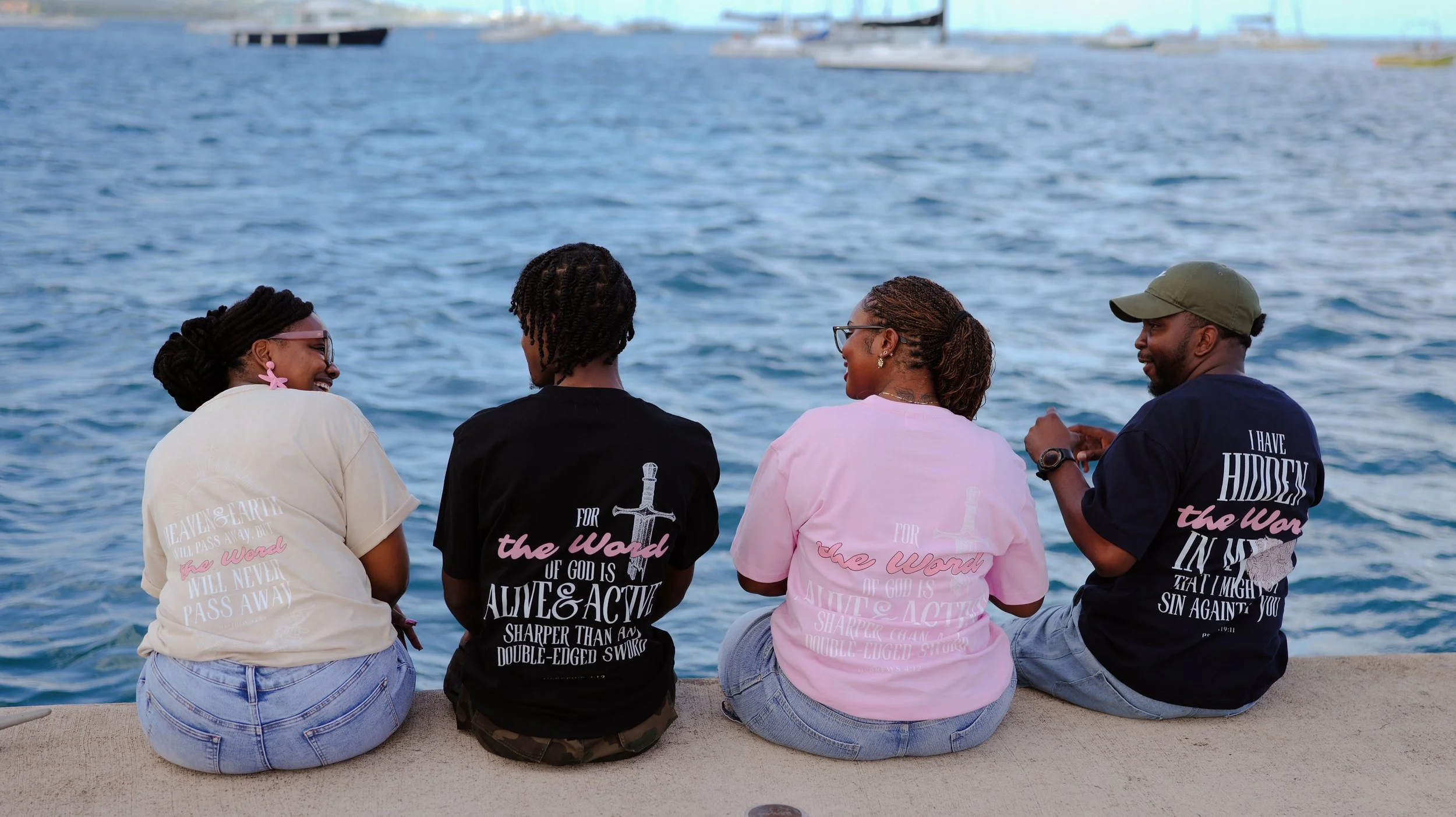 Four friends sitting by the water, facing away from the camera, wearing matching T-shirts with biblical messages, and enjoying a conversation.