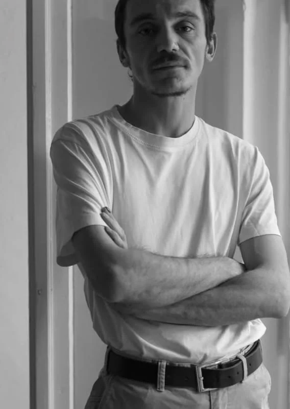 Black and white photo of a man standing with arms crossed, wearing a white T-shirt and light-colored pants, in front of a door.