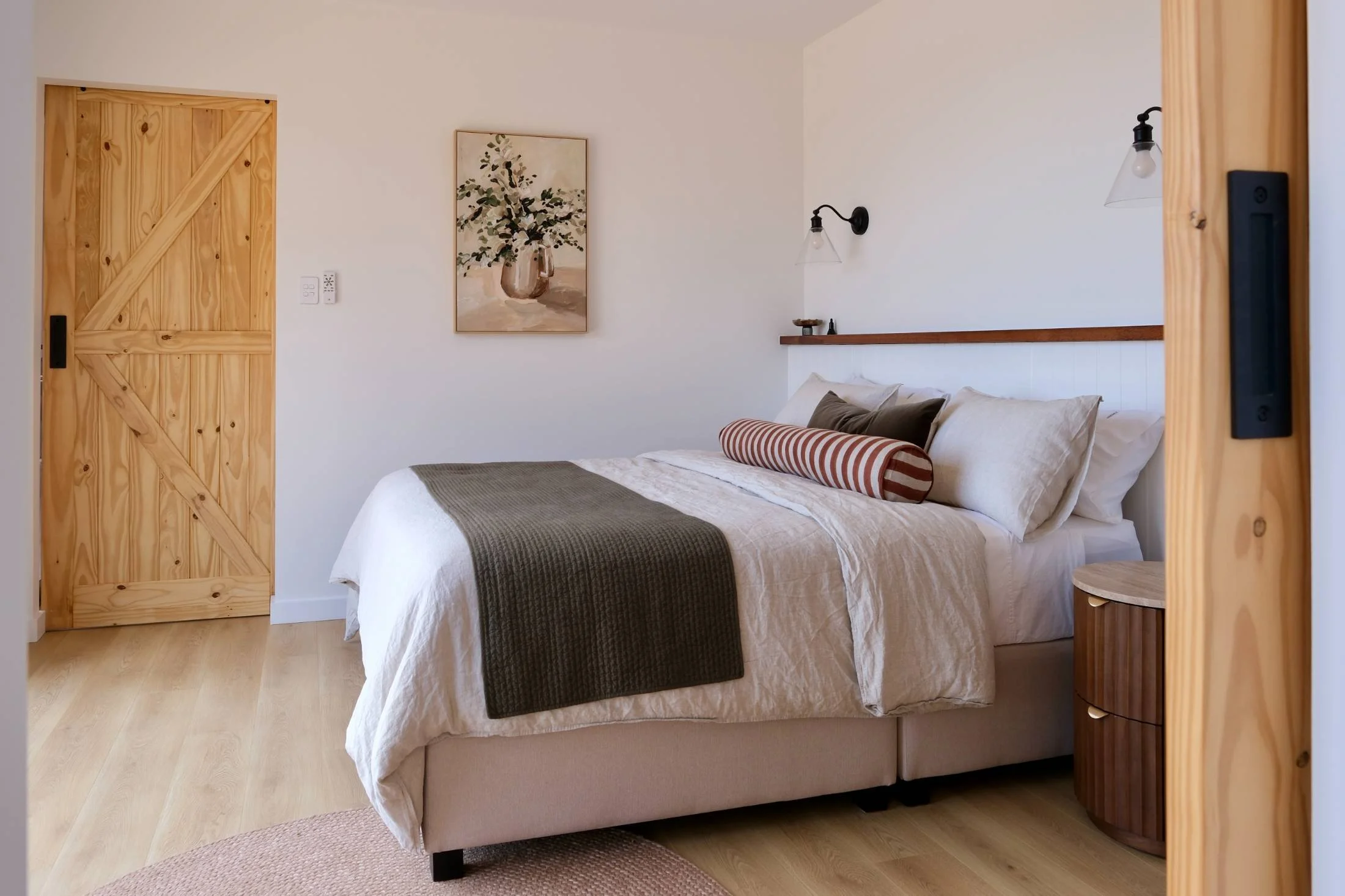 A cozy bedroom with a neatly made bed featuring white and beige linens, pillows, and a striped bolster pillow. The room has light wood flooring, a small wooden side table, wall-mounted black lamps, a painting of a vase with flowers, and a wooden slid
