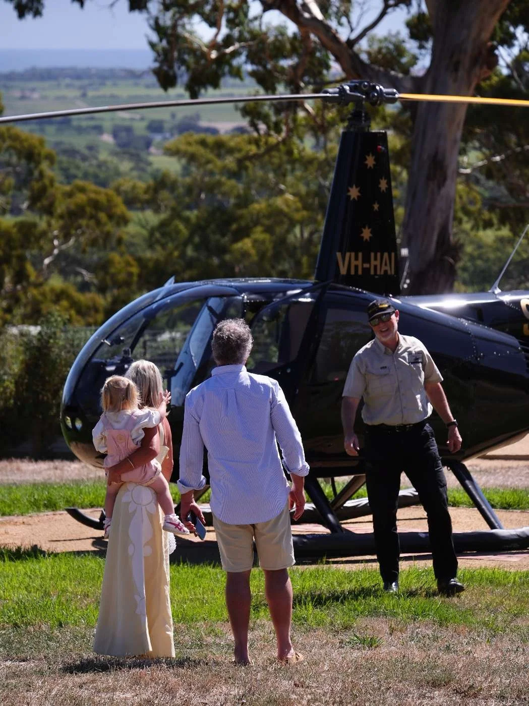 A family of three with a young girl, an older man, and a man in a pilot's uniform standing near a black helicopter outdoors, with trees and hills in the background.