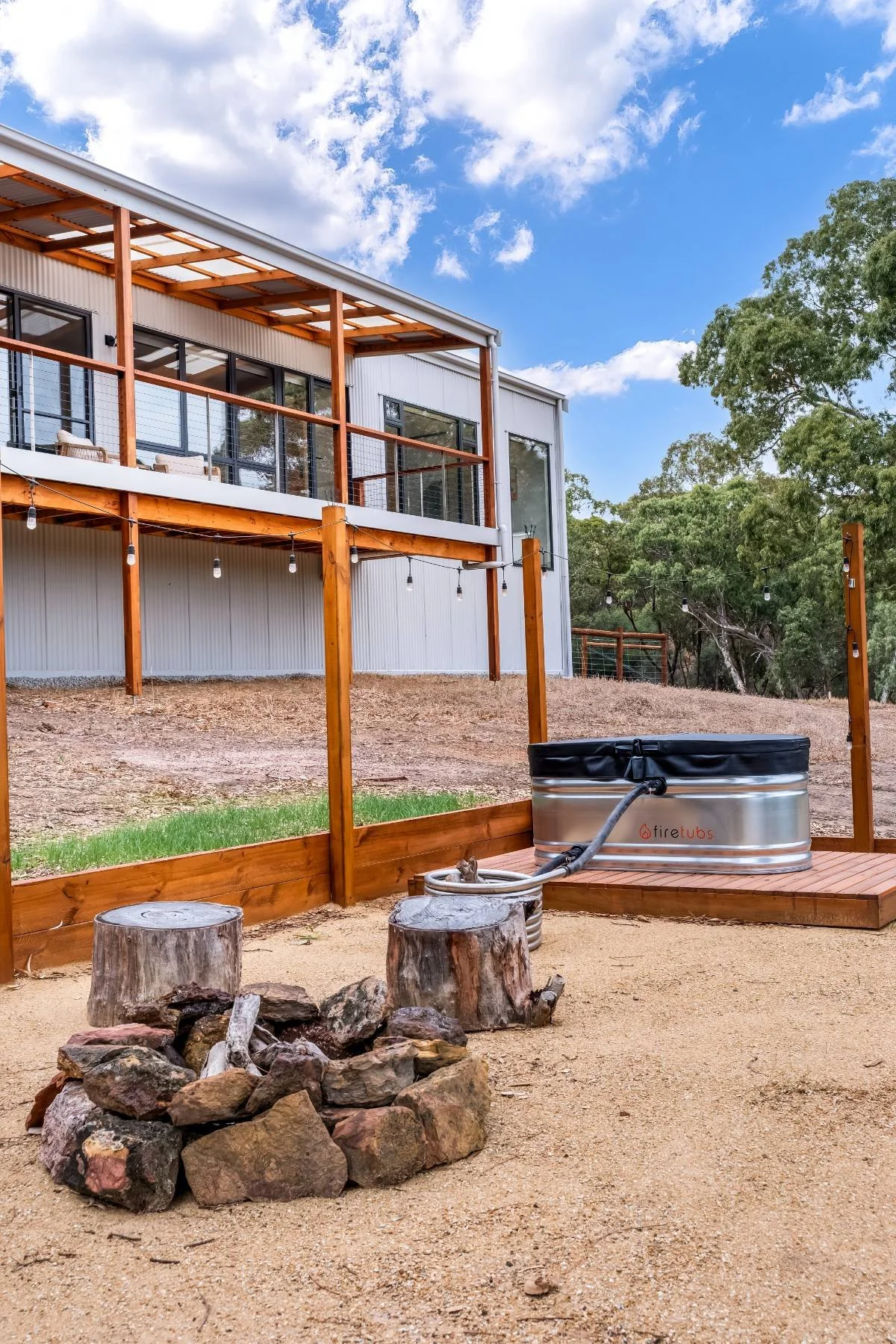 A modern two-story house with large windows and a spacious balcony. In front of the house, there is a fire tub with a hose connected, set on a wooden platform. A small fire pit made of rocks and two tree stumps are nearby on sandy ground. The backgro