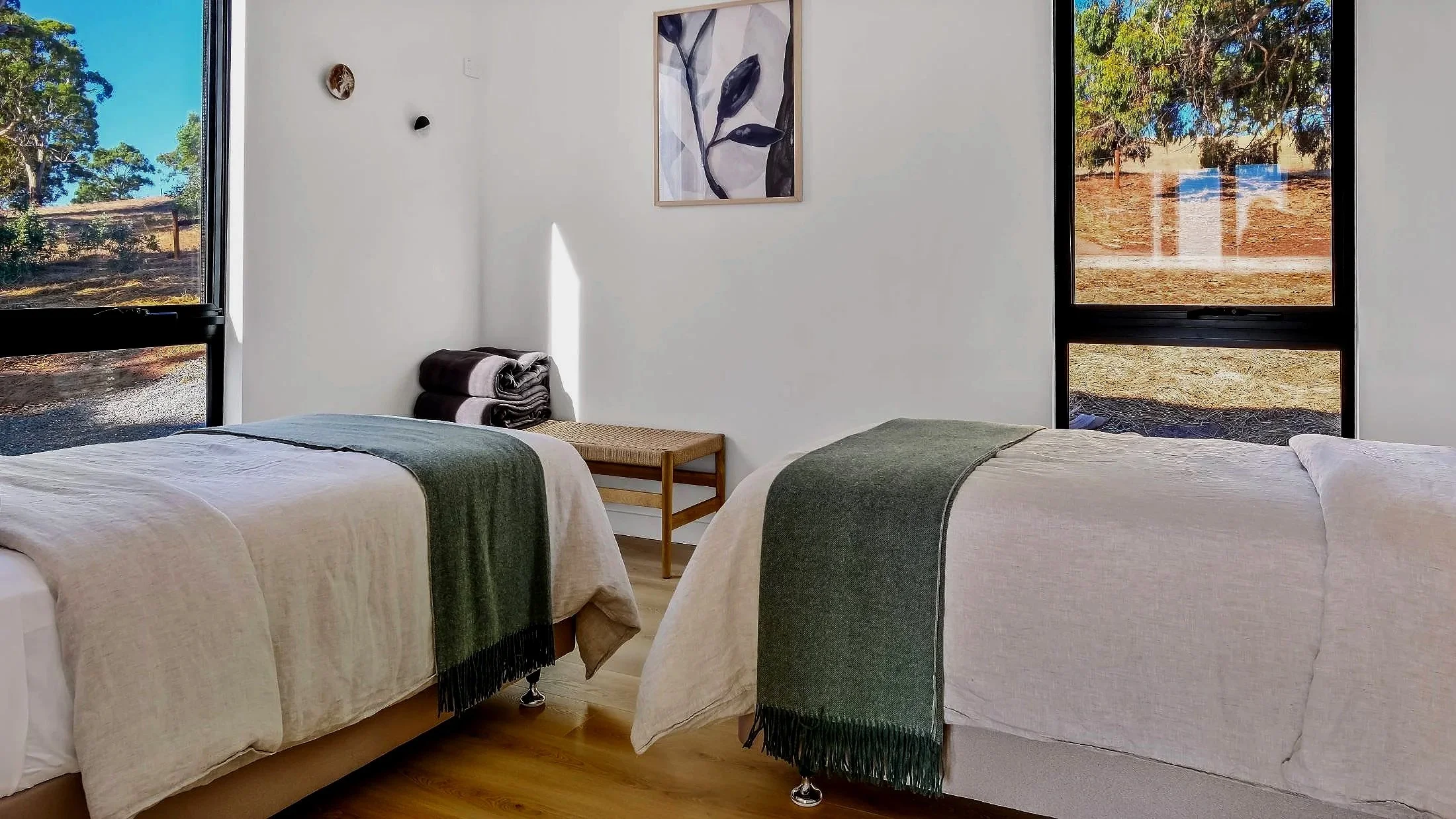 A bedroom with two single beds each with a green blanket, a small wooden bench, folded towels, and large windows showing an outdoor natural scene.