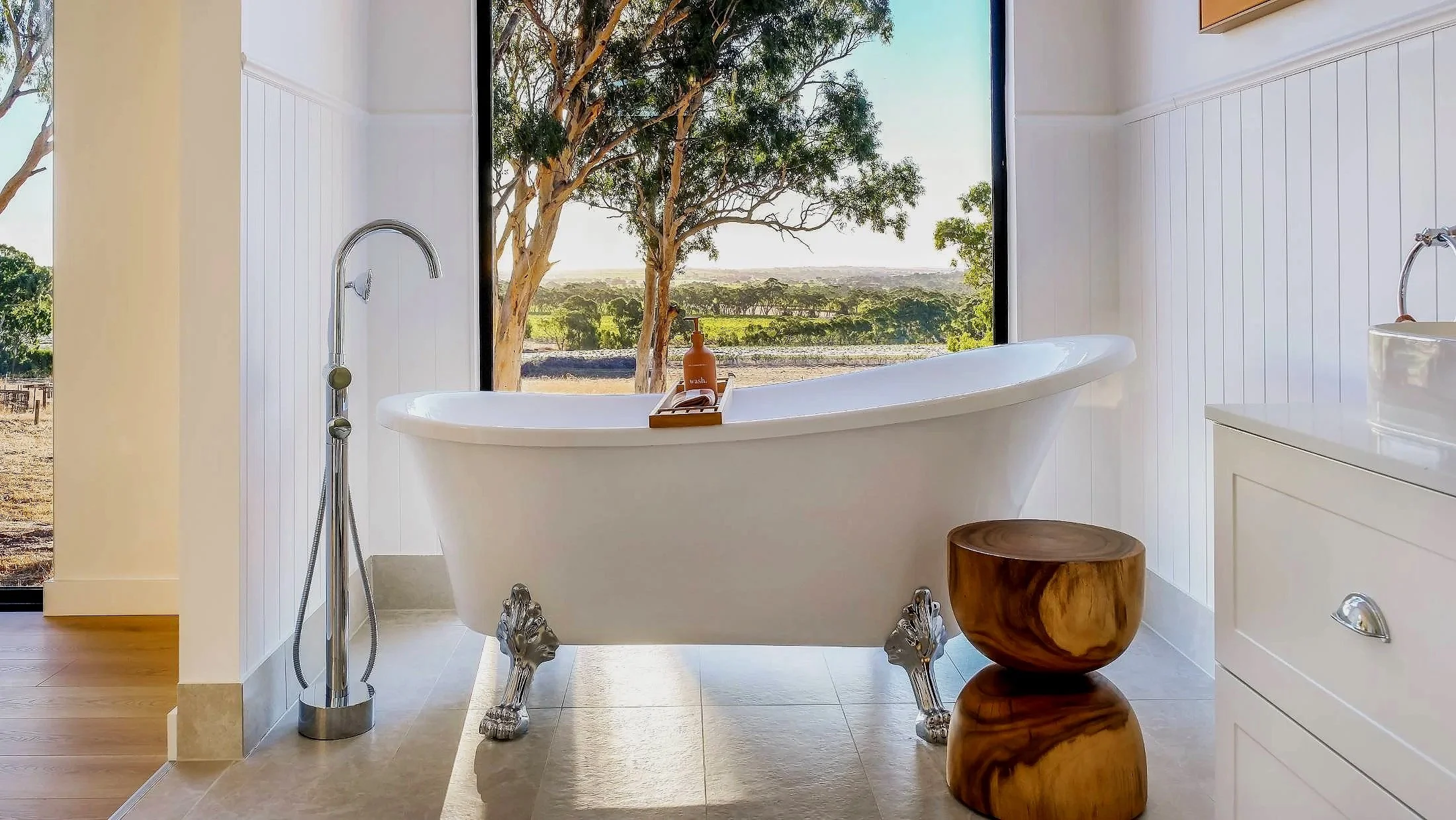 White clawfoot bathtub with silver lion head feet, placed in front of large window showing trees and a landscape, with a wooden side table beside it.