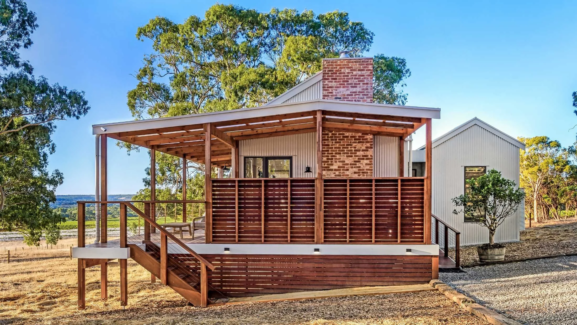 Modern house with a wooden deck and railing, white siding, brick chimney, surrounded by trees and open land, under a clear blue sky.