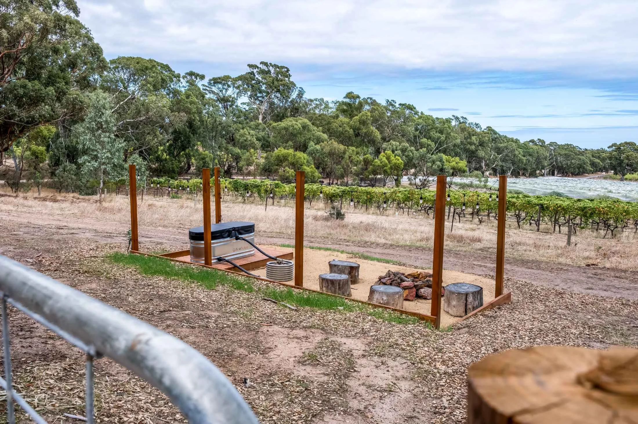 A backyard with a small fire pit and wooden stumps around it, enclosed by a wooden fence, overlooking a vineyard and a river in the background with trees and a cloudy sky.