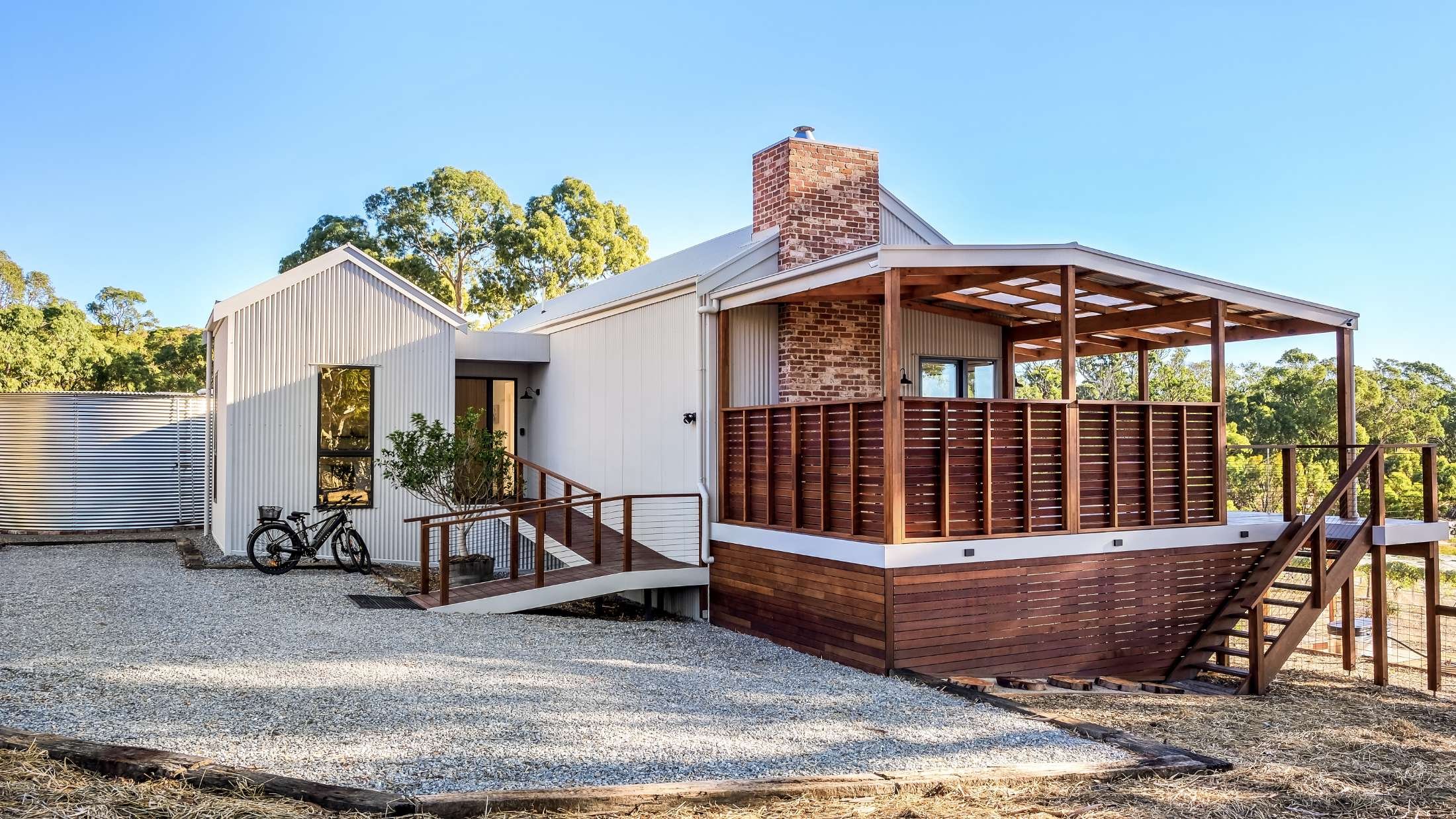 Modern house with metal siding and a wooden deck, outdoors during daytime, with trees in background and a gravel pathway in front.