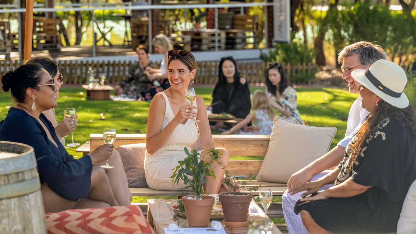 A group of people enjoying a daytime outdoor gathering, sitting on benches and chatting, with glasses of white wine, surrounded by greenery and plants.