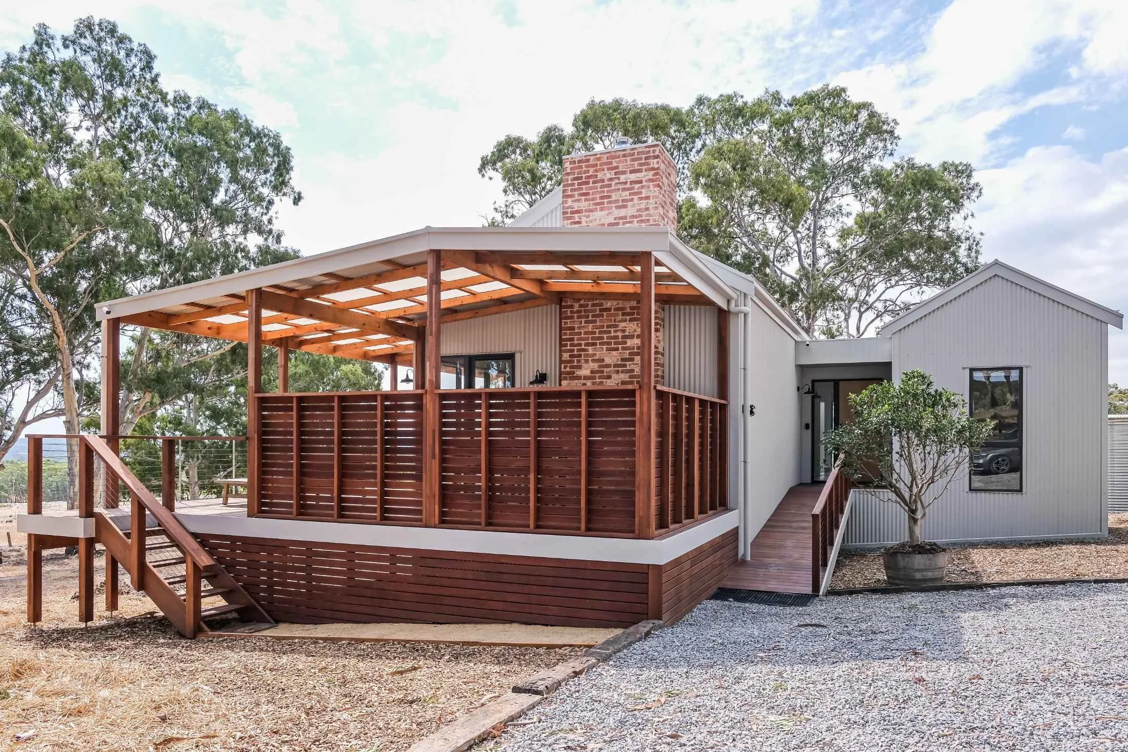 Modern house with a wooden deck and outdoor space, trees in the background, and a gravel ground.