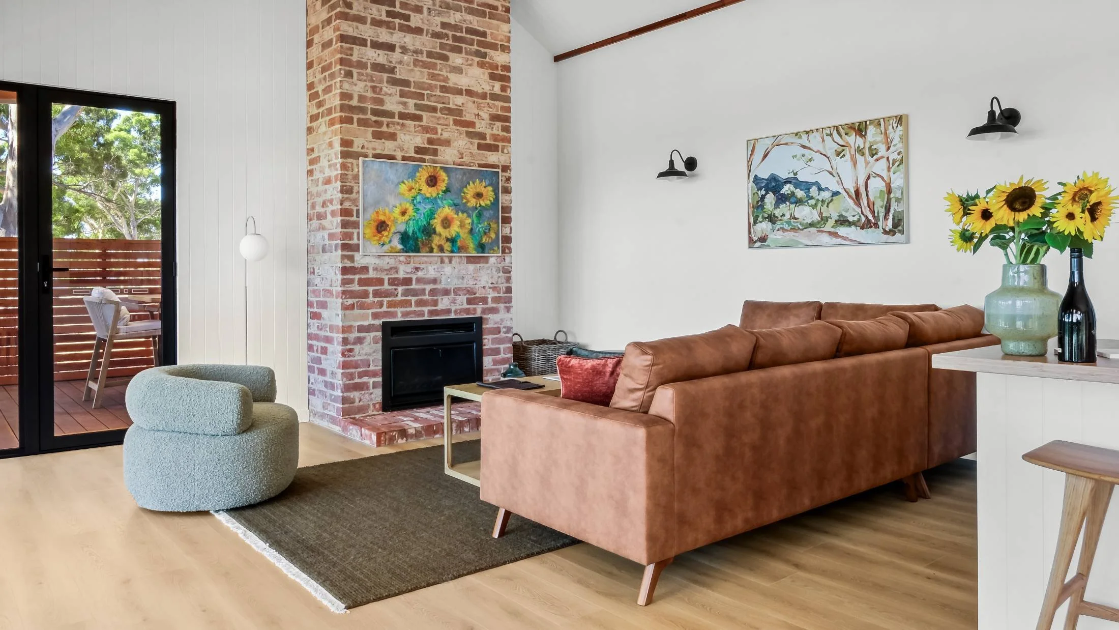 Living room with brick fireplace, wooden floors, brown couch, green armchair, wall art, sliding glass door to outdoor deck, flowers on side table.