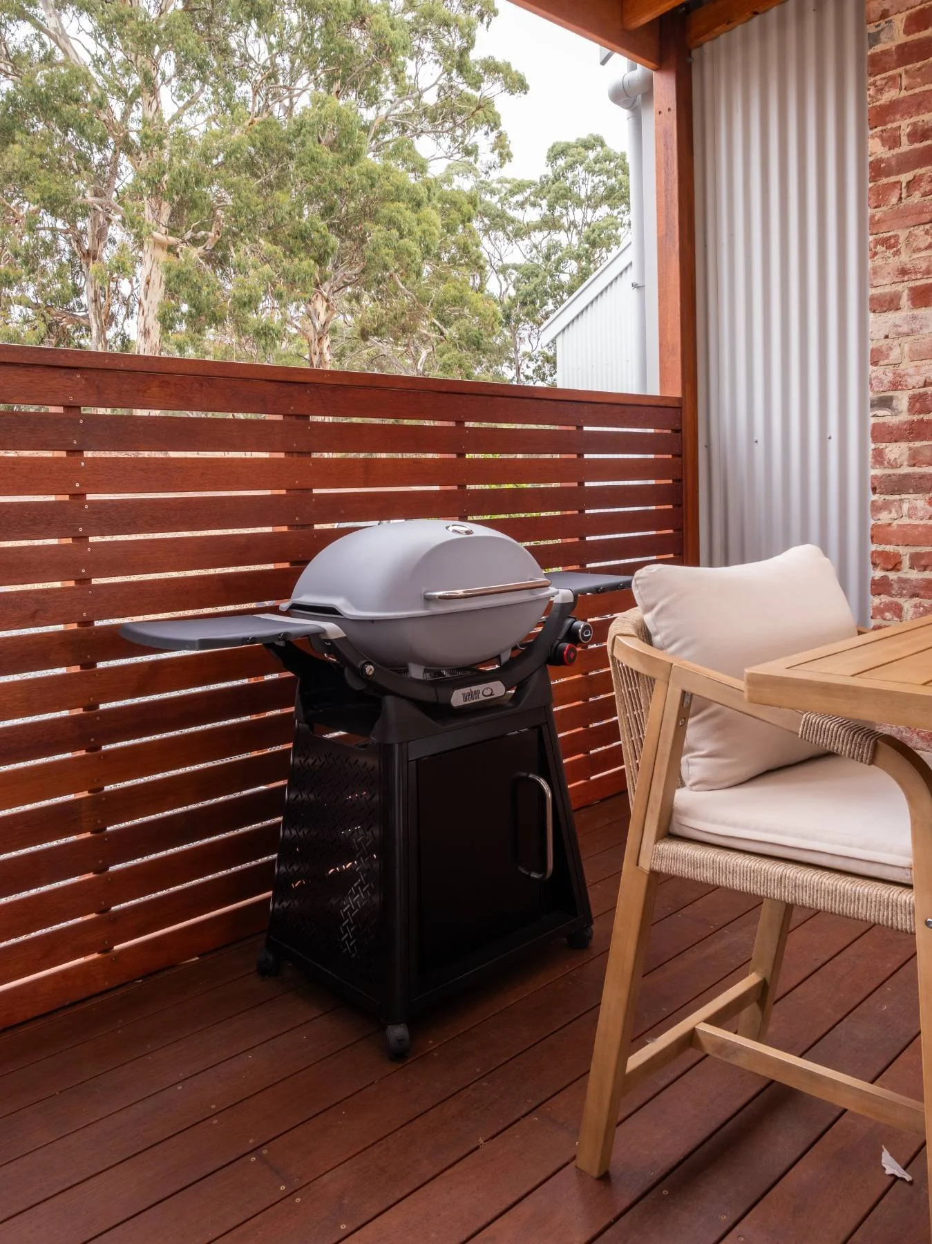 Outdoor balcony with wooden flooring, a wooden railing, a gray barbecue grill on a black stand, and a wooden chair with a white cushion.