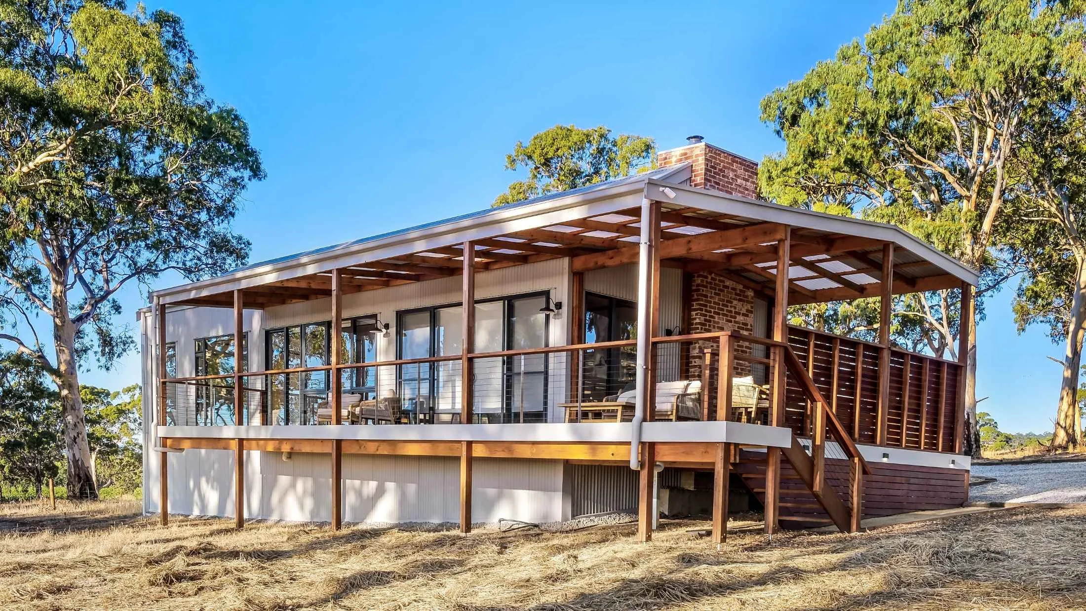 Modern house with wooden deck and railing, surrounded by trees, under a clear blue sky.