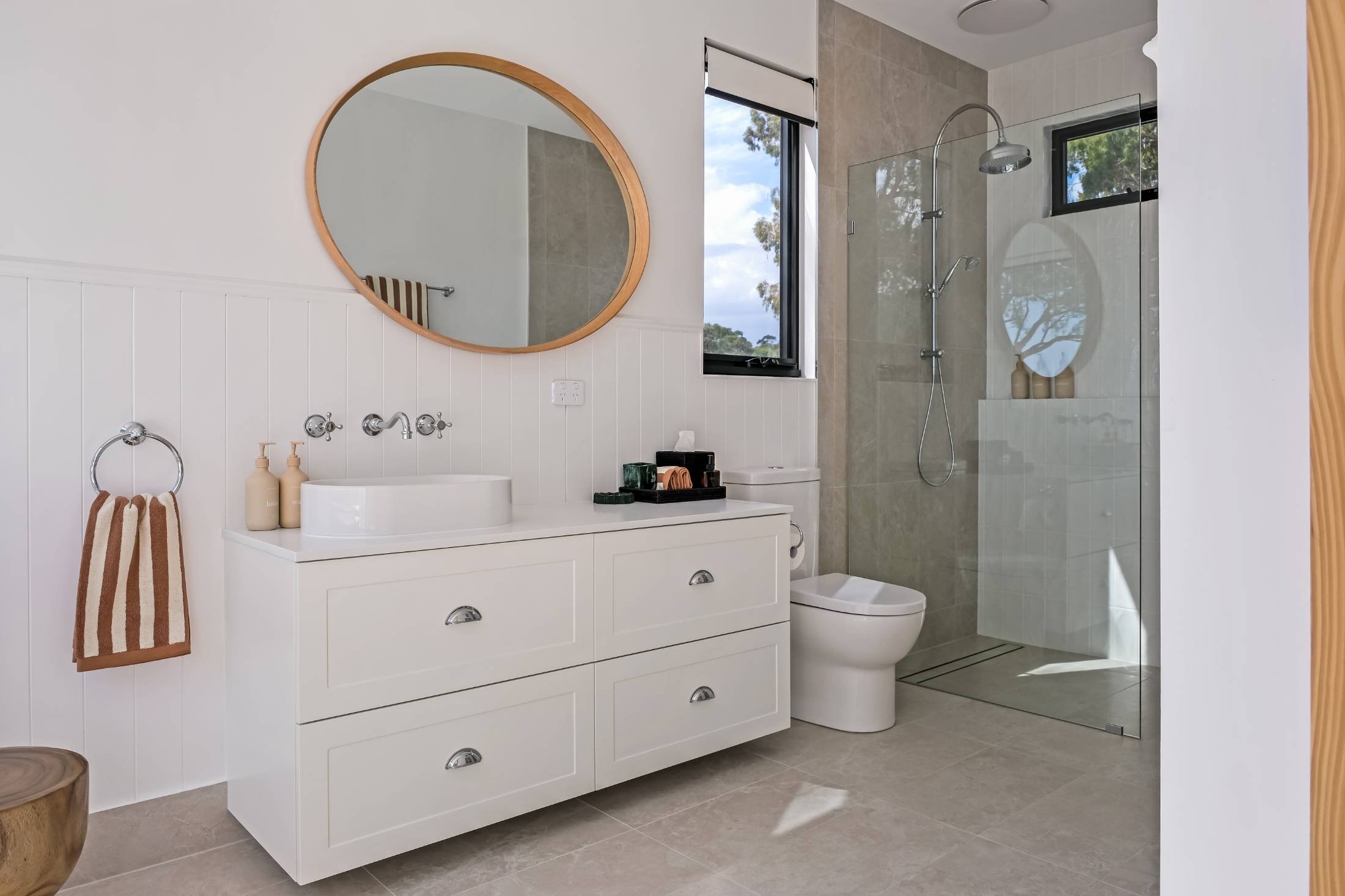 Modern bathroom with white vanity, oval mirror, beige and brown towels, toilet, and walk-in shower with glass door and window