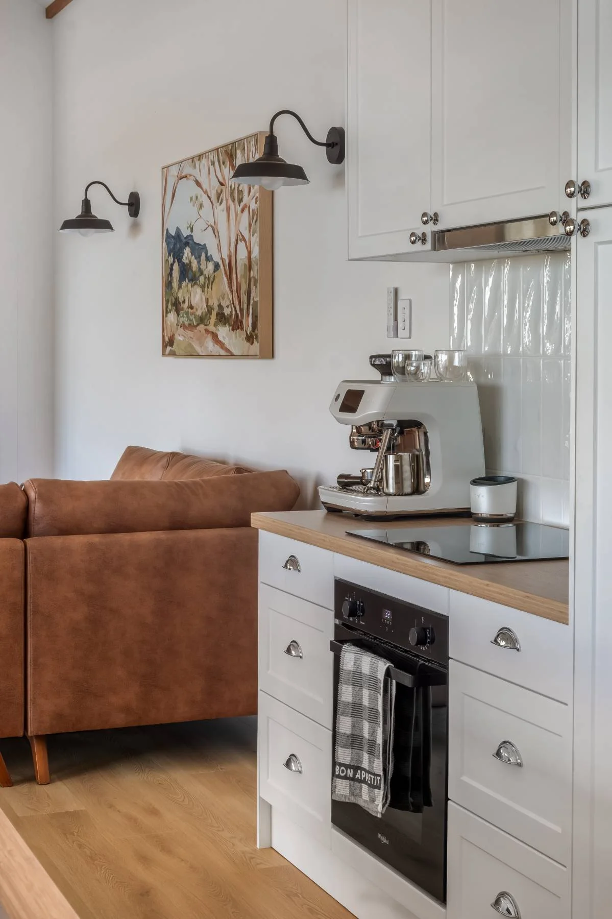 Modern kitchen with a white cabinet, coffee machine, and a black oven; next to a brown leather sofa; a painting and wall lights are also visible.