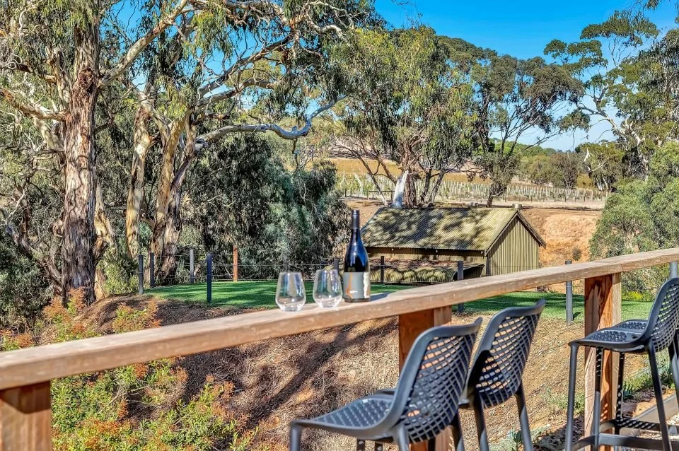 Outdoor patio with a wooden railing, three barstools, a wine bottle, and two wine glasses set on the railing, overlooking a landscape with trees, a small shed, and rolling hills under a clear blue sky.