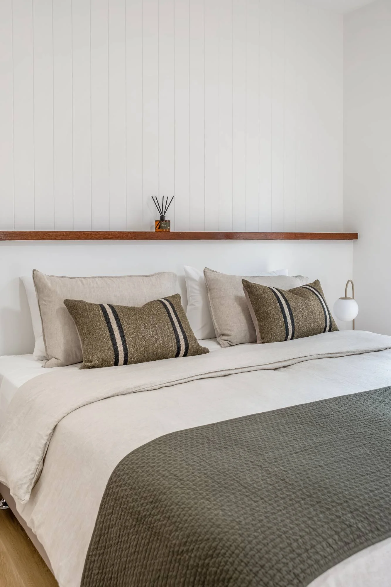 A neatly made bed with beige and taupe pillows, a textured taupe bedspread, against a white wall with a wooden shelf above it. On the shelf, there is a small diffuser and a small lamp with a round white shade.