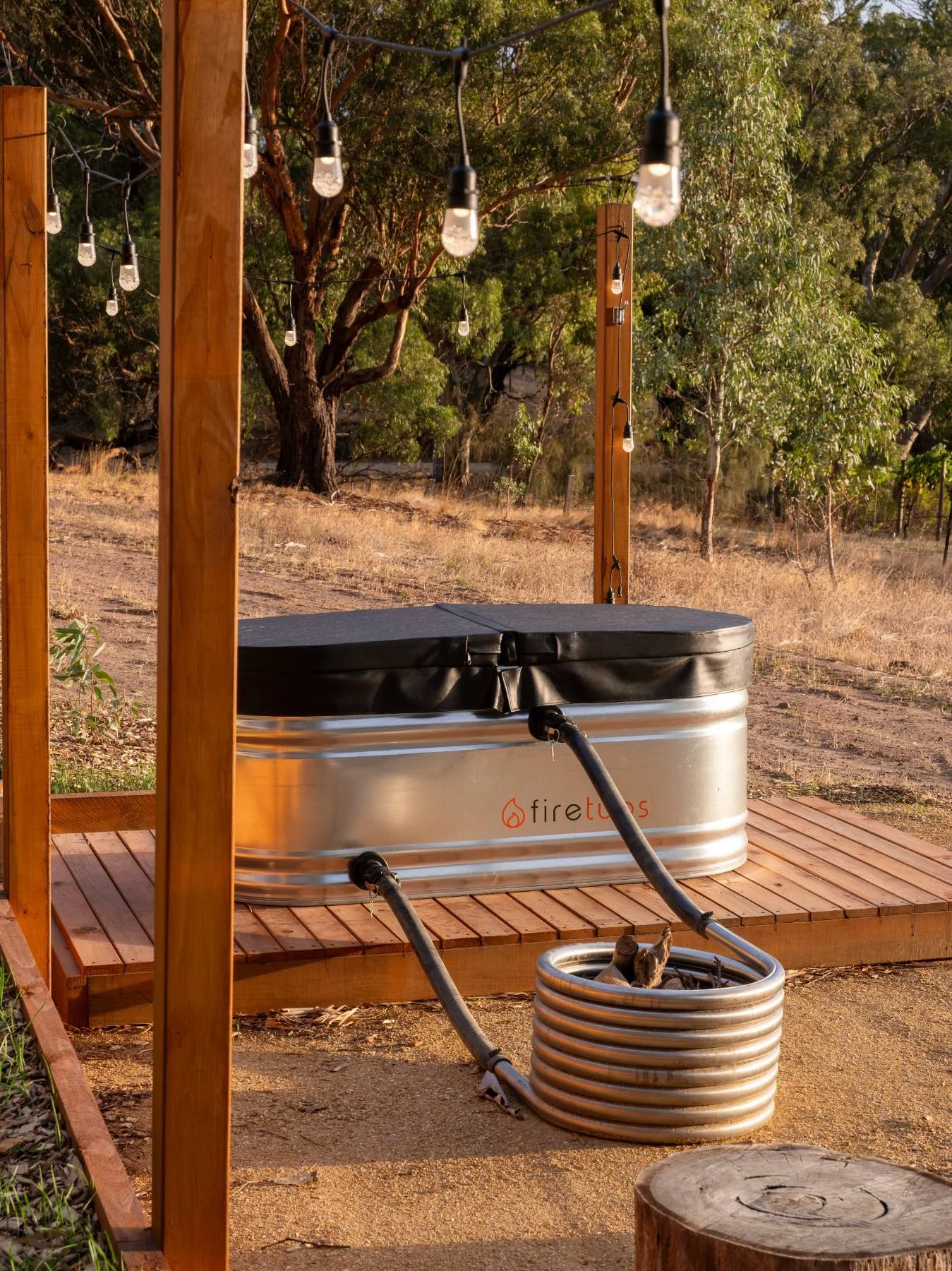 Outdoor hot tub with a metal exterior and black cover, on a wooden platform, under string lights, in a natural setting with trees and dry grass.