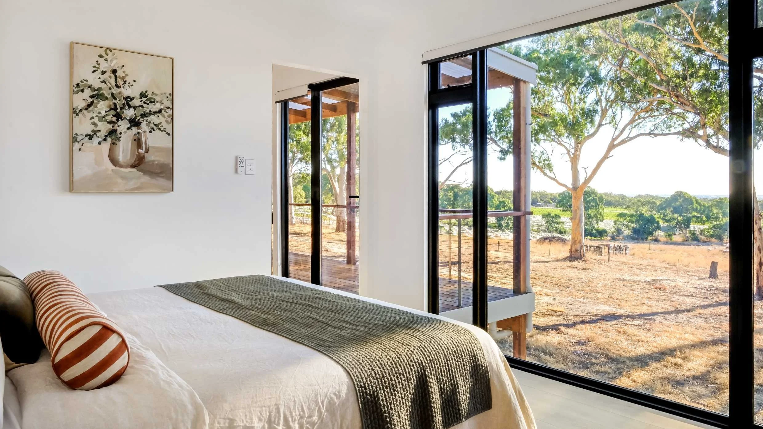 A bedroom with large floor-to-ceiling windows showing an outdoor view of trees and a dry landscape. There is a bed with white sheets, a green blanket, and a striped pillow. A painting of a vase with flowers hangs on the wall.