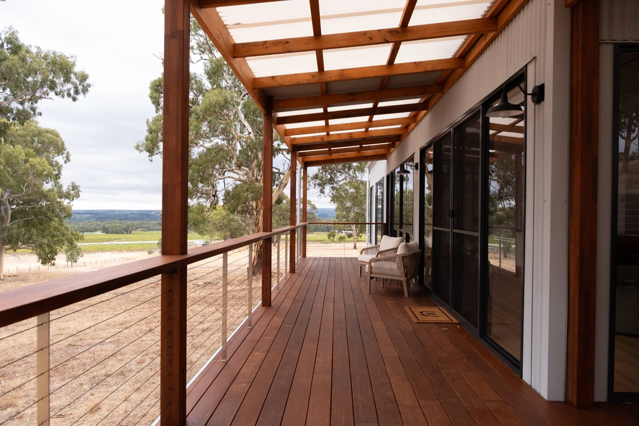 A wooden balcony with a railing overlooking a rural landscape of trees and fields, with a sliding glass door leading into a building.