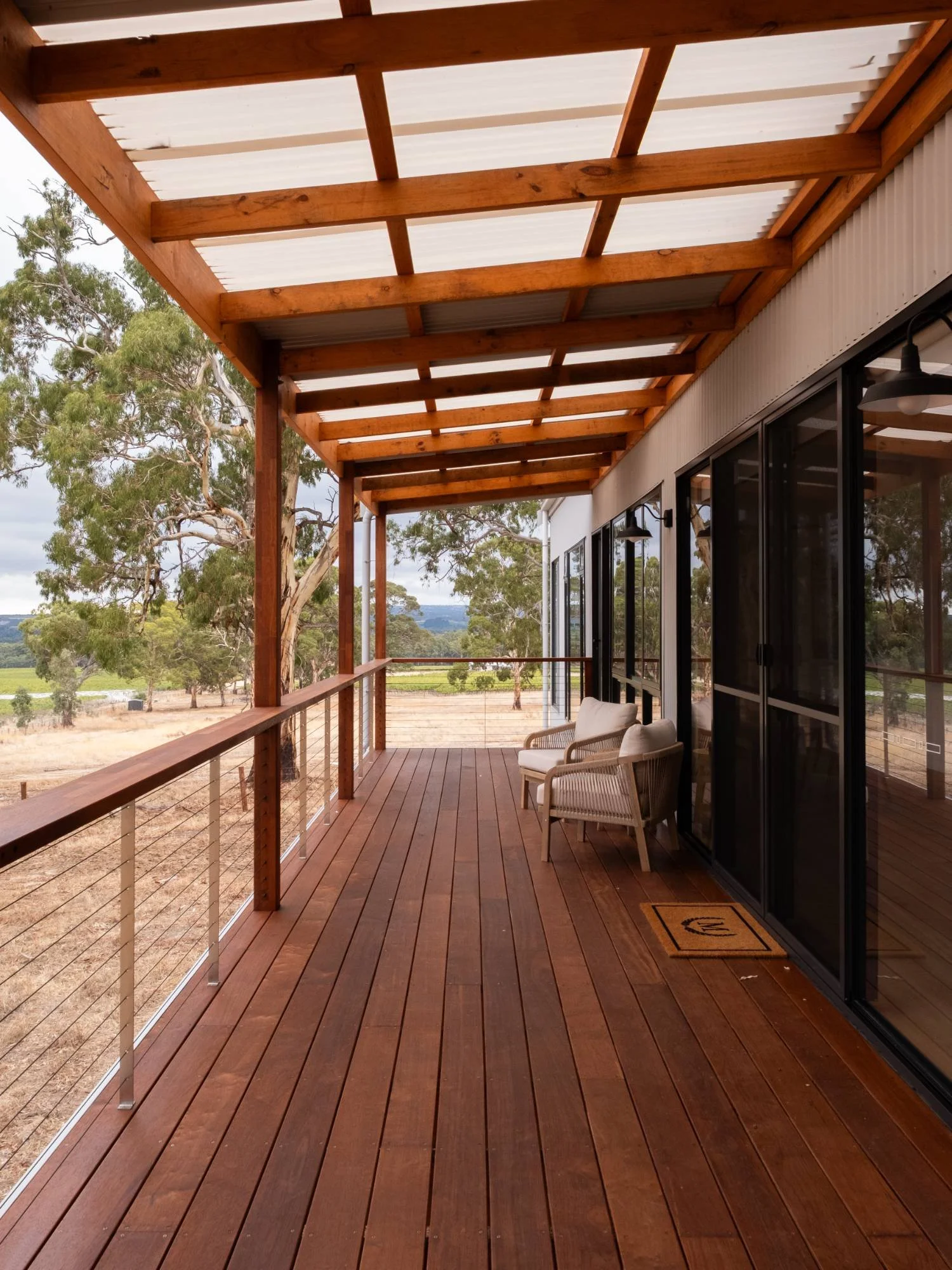 A wooden outdoor balcony with a railing, two chairs, and a doormat, overlooking a landscape with trees and open fields under a cloudy sky.